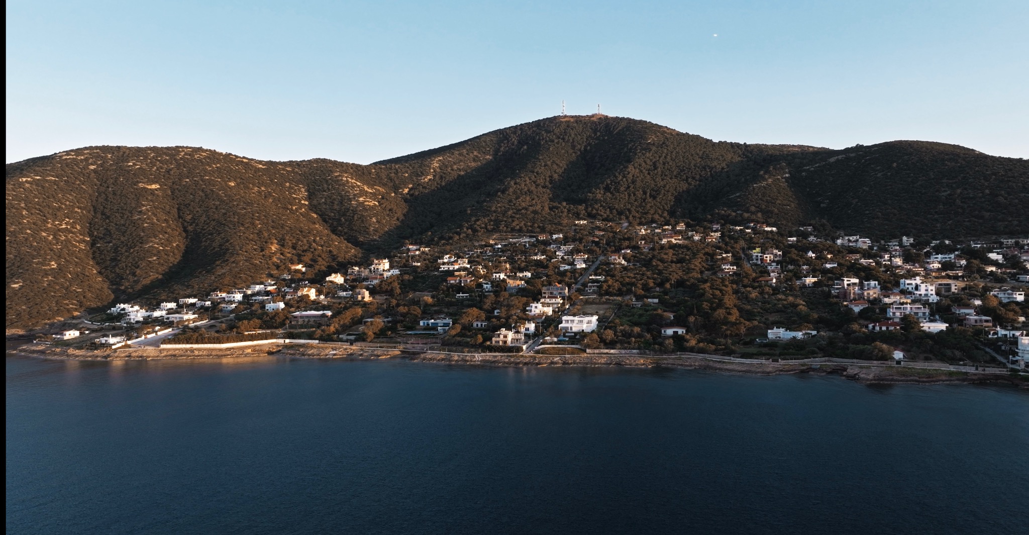 Zdjęcie z lotu ptaka przedstawia nadmorskie miasteczko z białymi domami na zboczu góry, z anteną na szczycie. W dole ciemna woda i błękitne niebo.