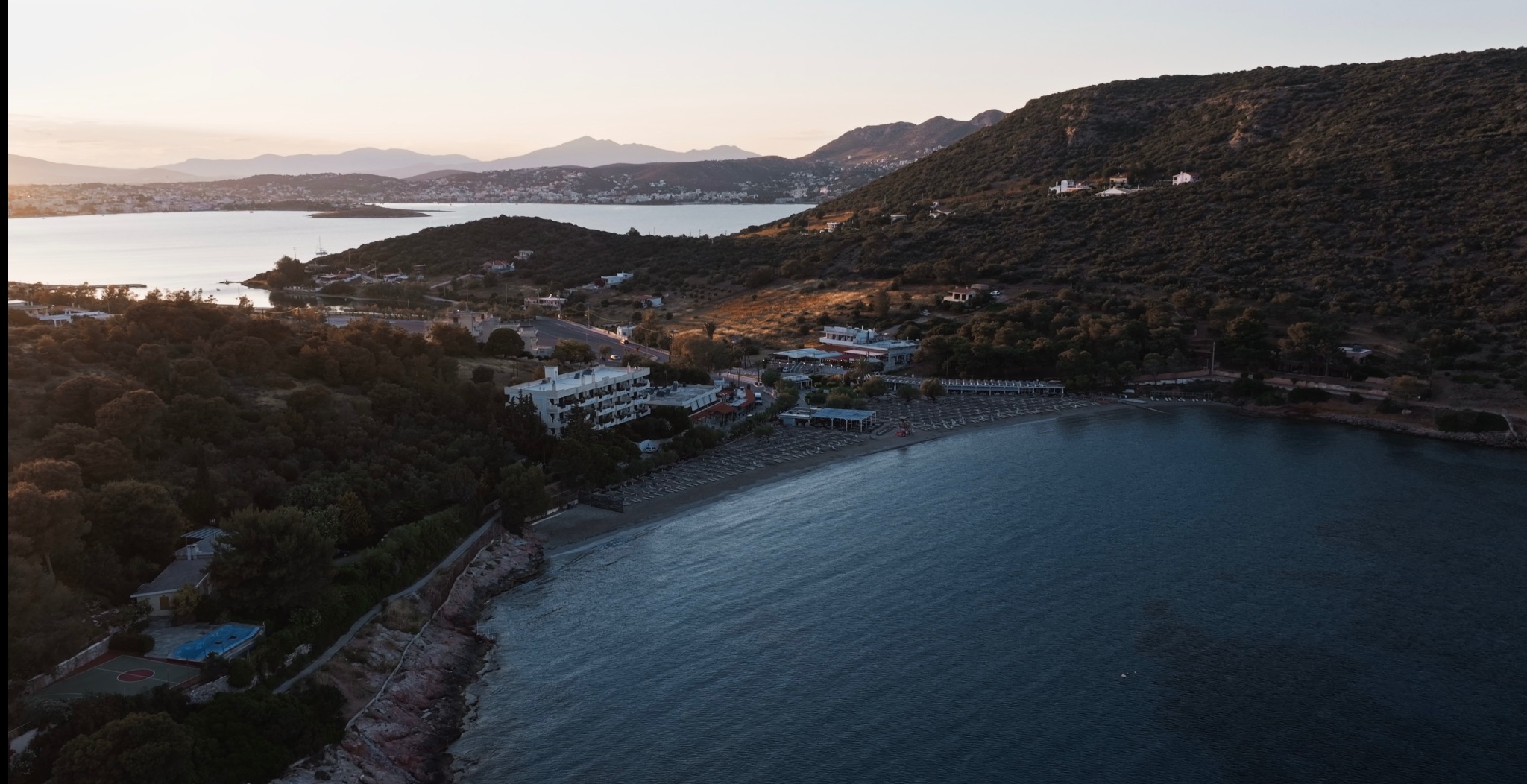 Panoramiczny widok z lotu ptaka na nadmorski kurort z plażą, hotelem i gęstą roślinnością w tle, uchwycony w miękkim świetle wczesnego ranka lub późnego popołudnia.