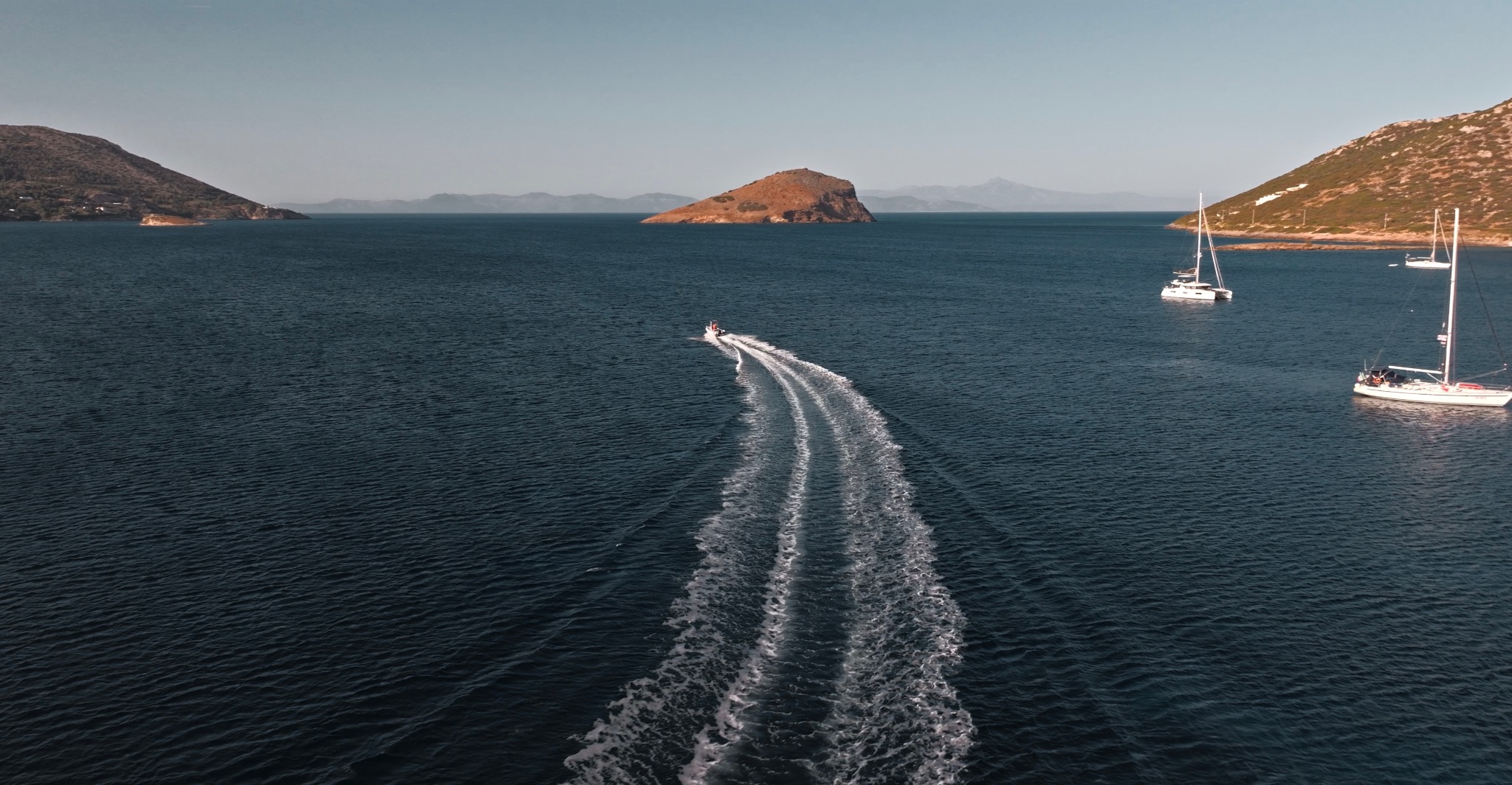 Błękitne morze z motorówką zostawiającą ślad piany, w tle górzyste wyspy i żaglówki, słoneczny dzień.