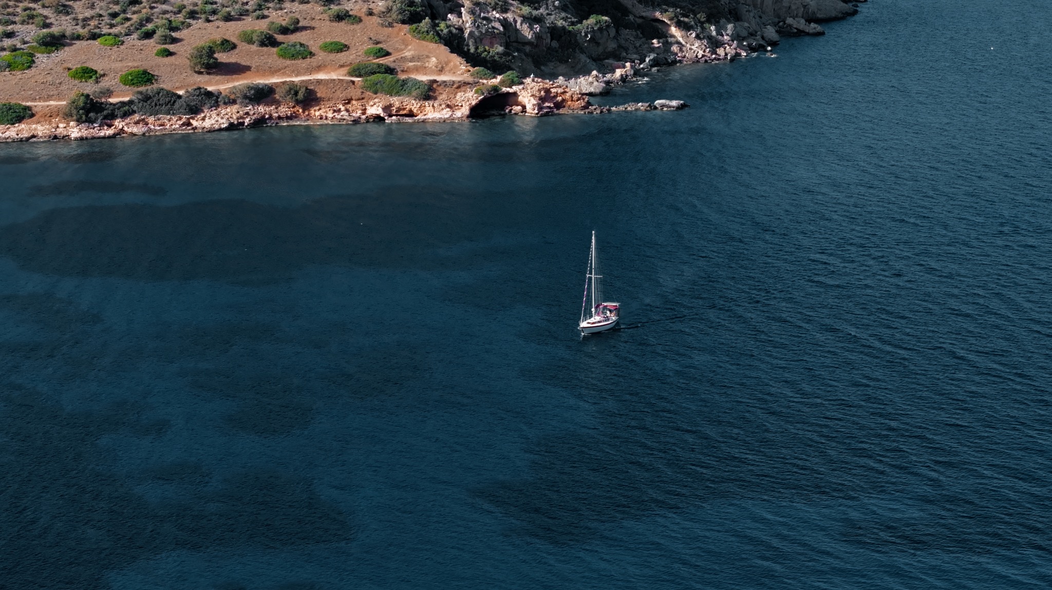 Błękitne morze z jachtem żaglowym płynącym wzdłuż skalistego brzegu z niską roślinnością. Ujęcie z góry, podkreślające głębię koloru wody i kontrast z lądem.
