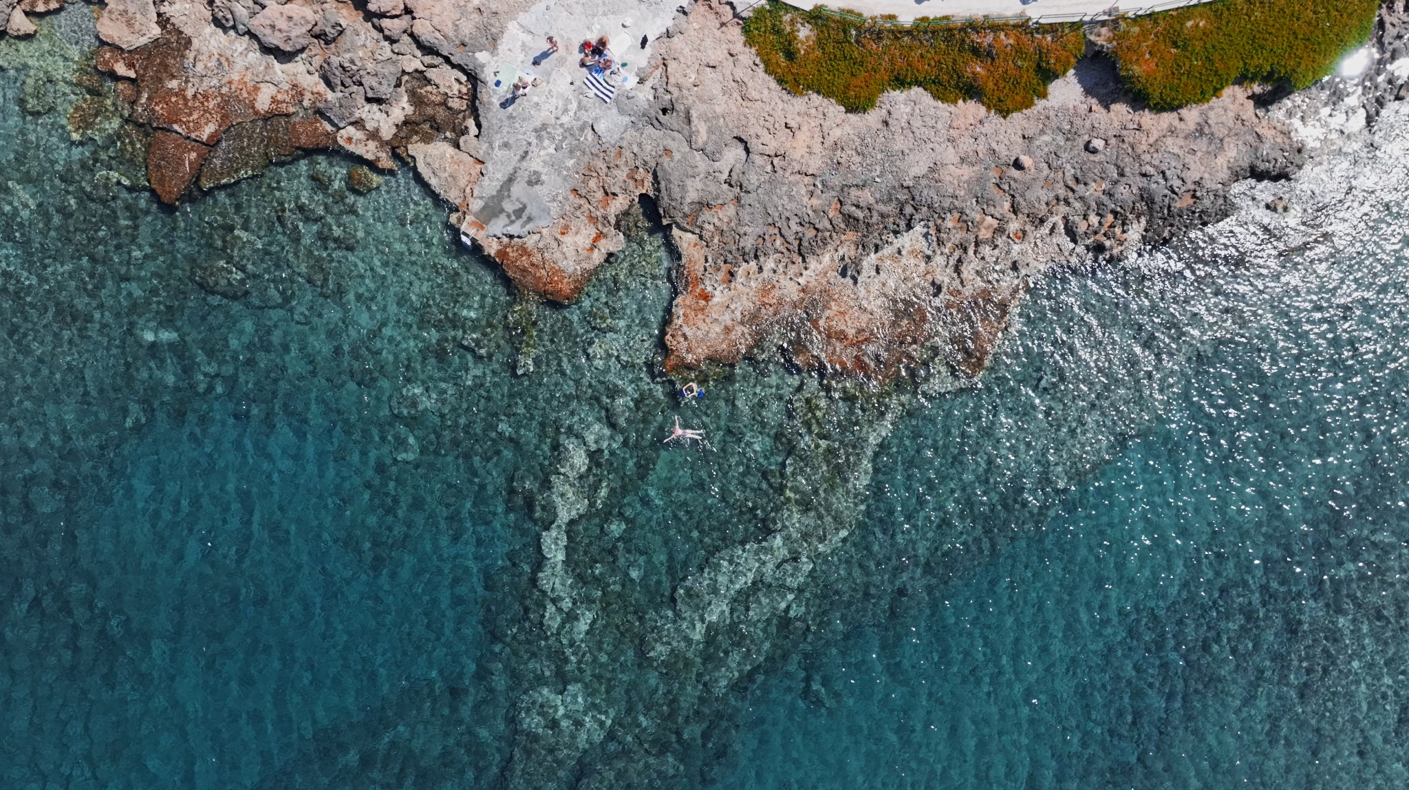 Widok z góry na krystalicznie czystą wodę obmywającą skalisty brzeg z osobami odpoczywającymi na ręczniku i pływającą osobą, uchwycony w słoneczny dzień.