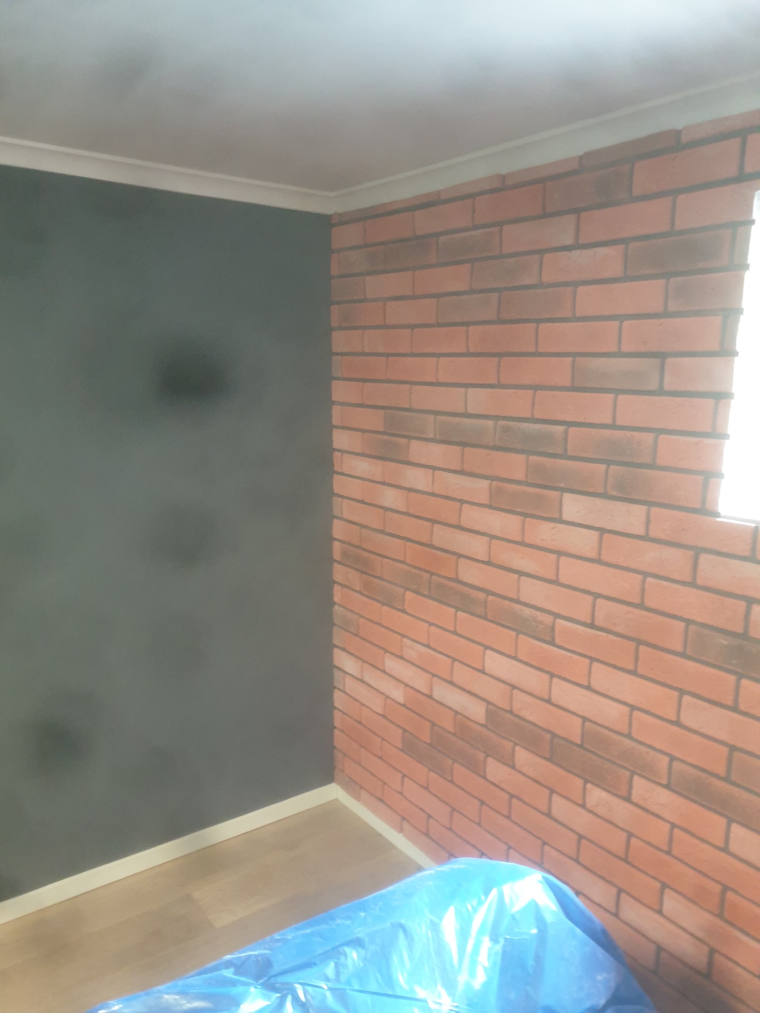 Pokój po remoncie: pomalowana na szaro ściana i ściana wyłożona czerwoną cegłą. Podłoga zabezpieczona folią. Widoczne listwy przypodłogowe i sufitowe.