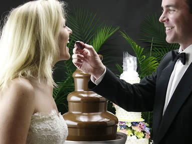 Para młoda podczas wesela korzysta z fontanny czekoladowej. Mężczyzna w smokingu podaje kobiecie w sukni ślubnej owoc zanurzony w czekoladzie, w tle tort weselny i zielone rośliny.