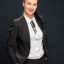 Portret kobiety w czarnym garniturze i białej koszuli z kokardą, uśmiechniętej i patrzącej w obiektyw na ciemnym tle.