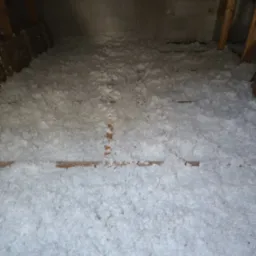 Poddasze z widoczną warstwą izolacji z białej wełny mineralnej, ułożoną między drewnianymi belkami, na tle ściany z białych bloczków betonowych.