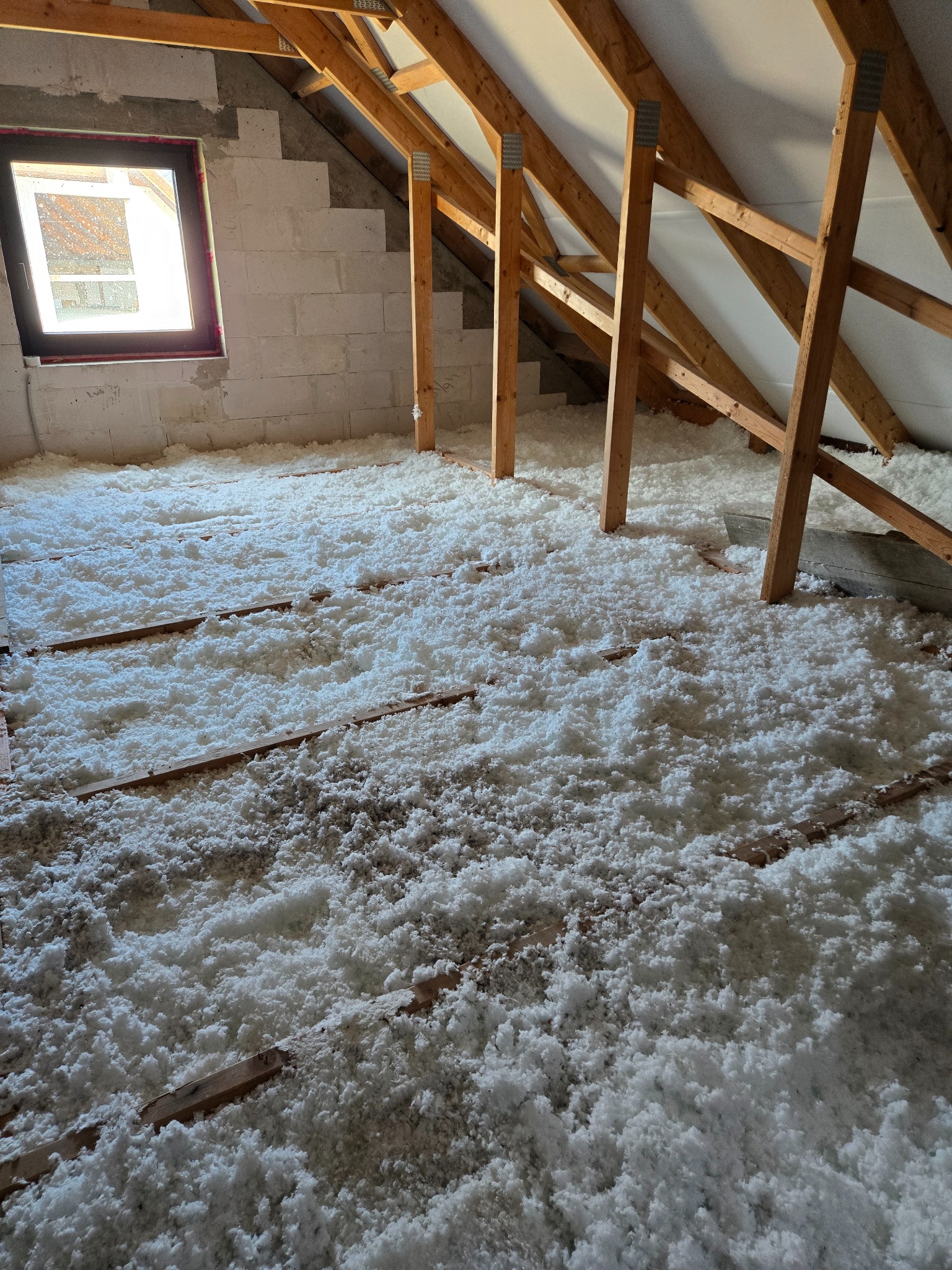 Poddasze z drewnianą konstrukcją dachu, częściowo ocieplone białą wełną mineralną. Widoczne bloczki betonowe i okno w szczytowej ścianie.