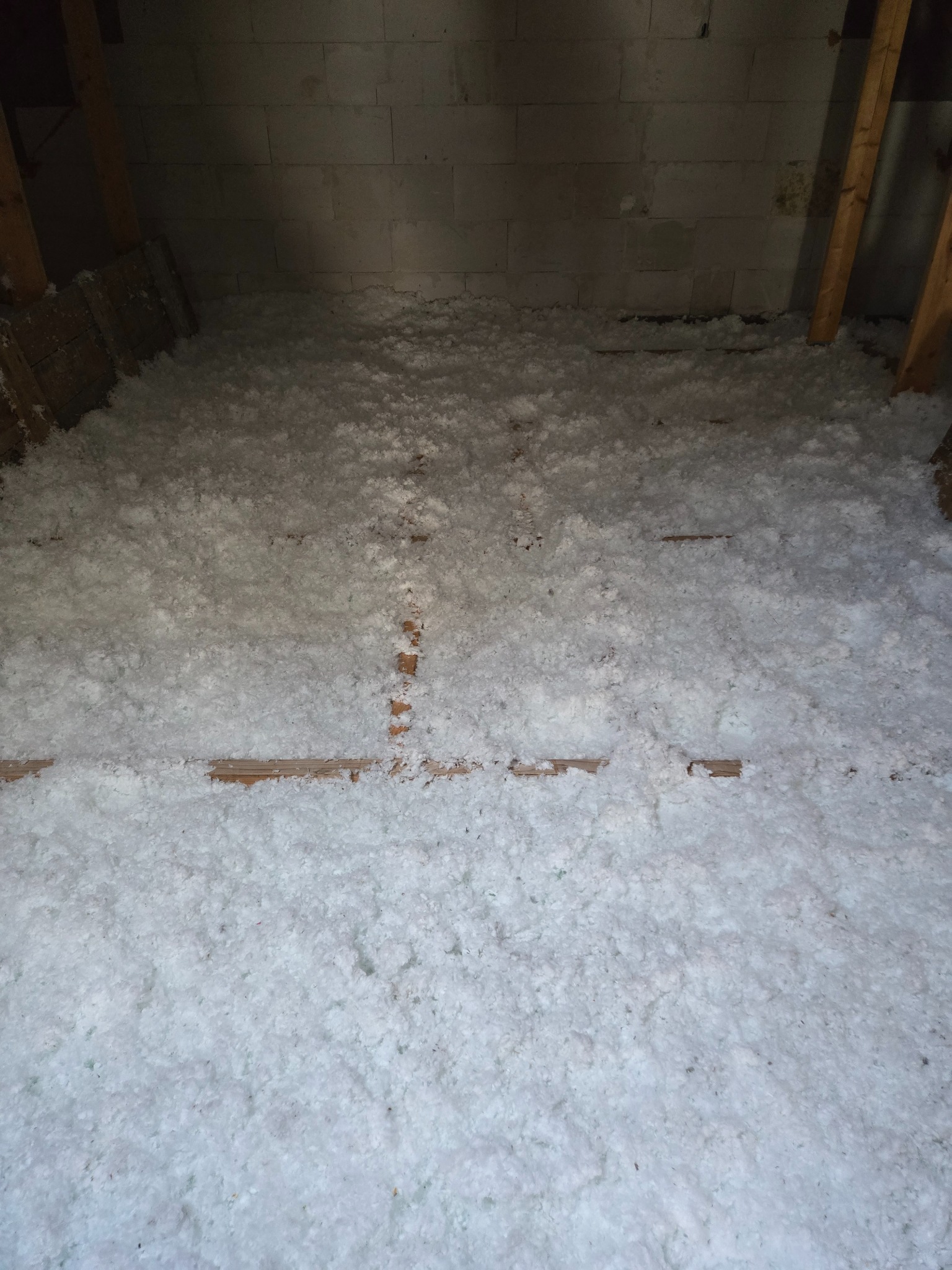 Poddasze z widoczną warstwą izolacji z białej wełny mineralnej, ułożoną między drewnianymi belkami, na tle ściany z białych bloczków betonowych.