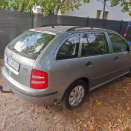 Szara Skoda Fabia Combi z hakiem holowniczym zaparkowana na żwirowym podłożu, z widocznym niebieskim busem w tle. Widok z tyłu i boku.