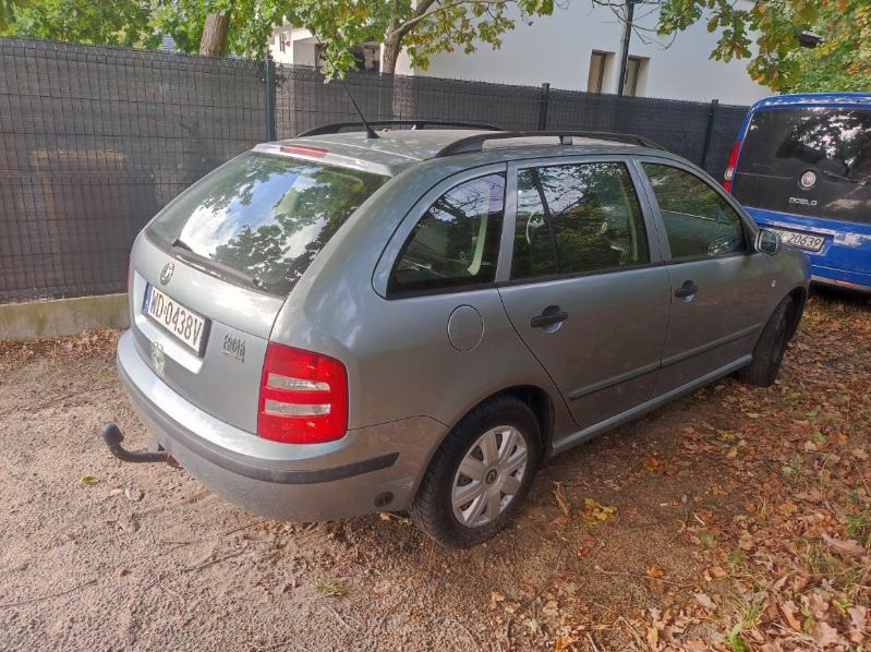 Szara Skoda Fabia Combi z hakiem holowniczym zaparkowana na żwirowym podłożu, z widocznym niebieskim busem w tle. Widok z tyłu i boku.