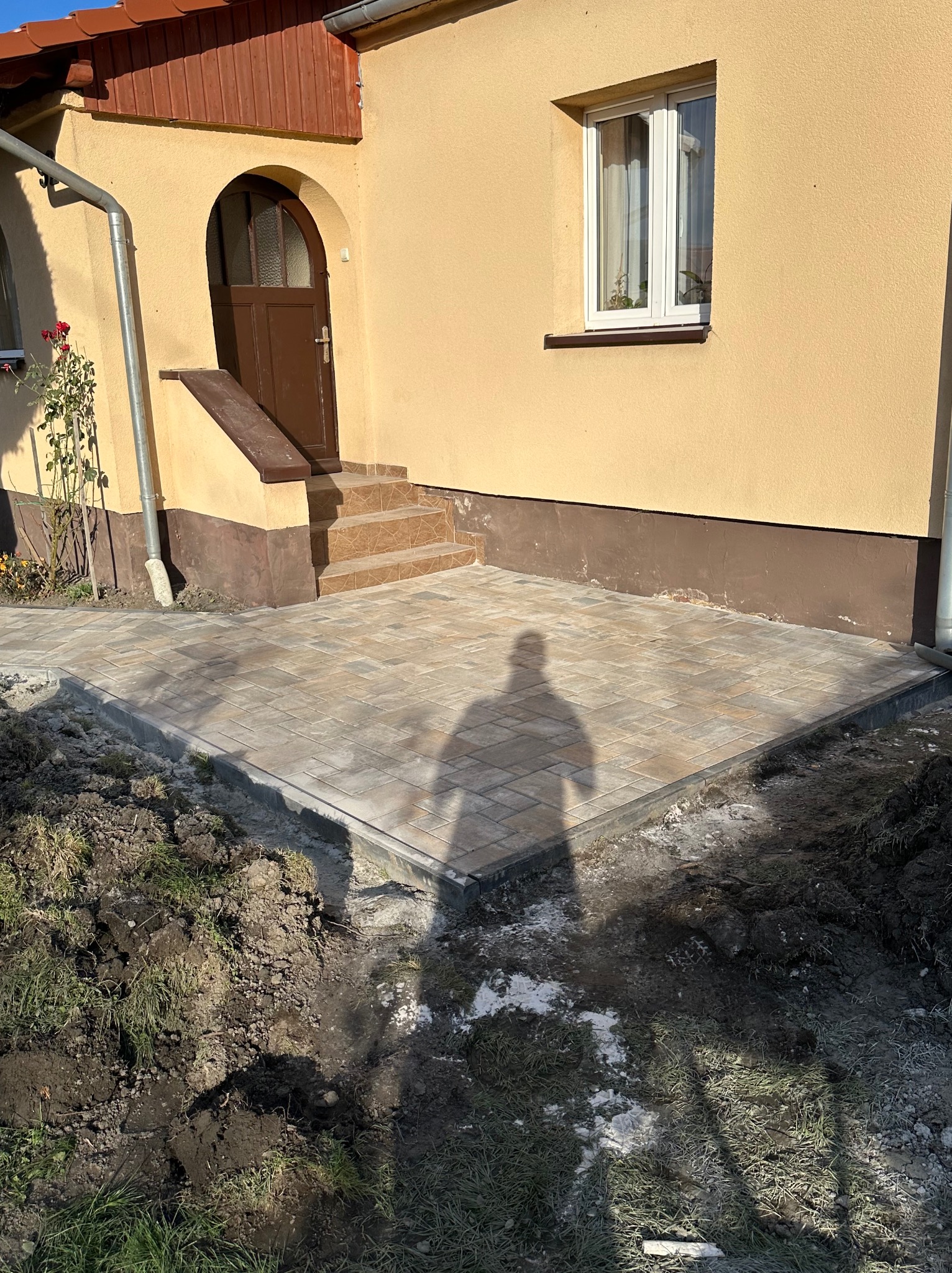 Ułożona kostka brukowa przed wejściem do domu z brązowymi drzwiami i schodami. Cień osoby na kostce. Prace ziemne w toku, widoczna ziemia i trawa.