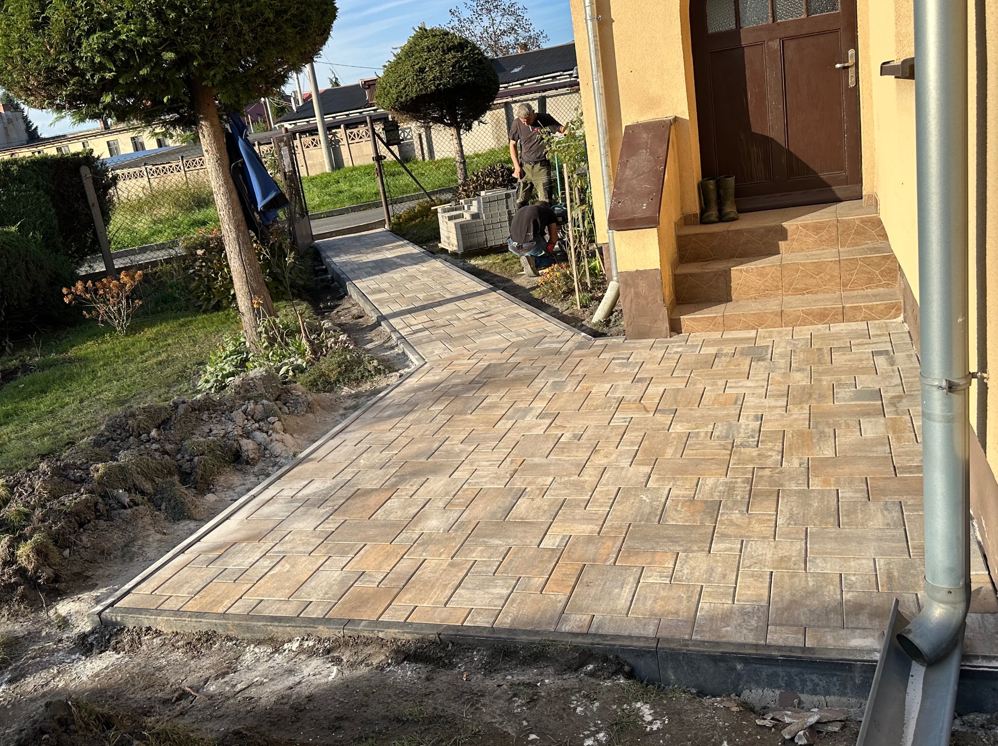 Ułożona kostka brukowa tworząca ścieżkę prowadzącą do domu. Widoczni pracownicy podczas układania kostki. Jasna, beżowo-brązowa kostka, szara obwódka.