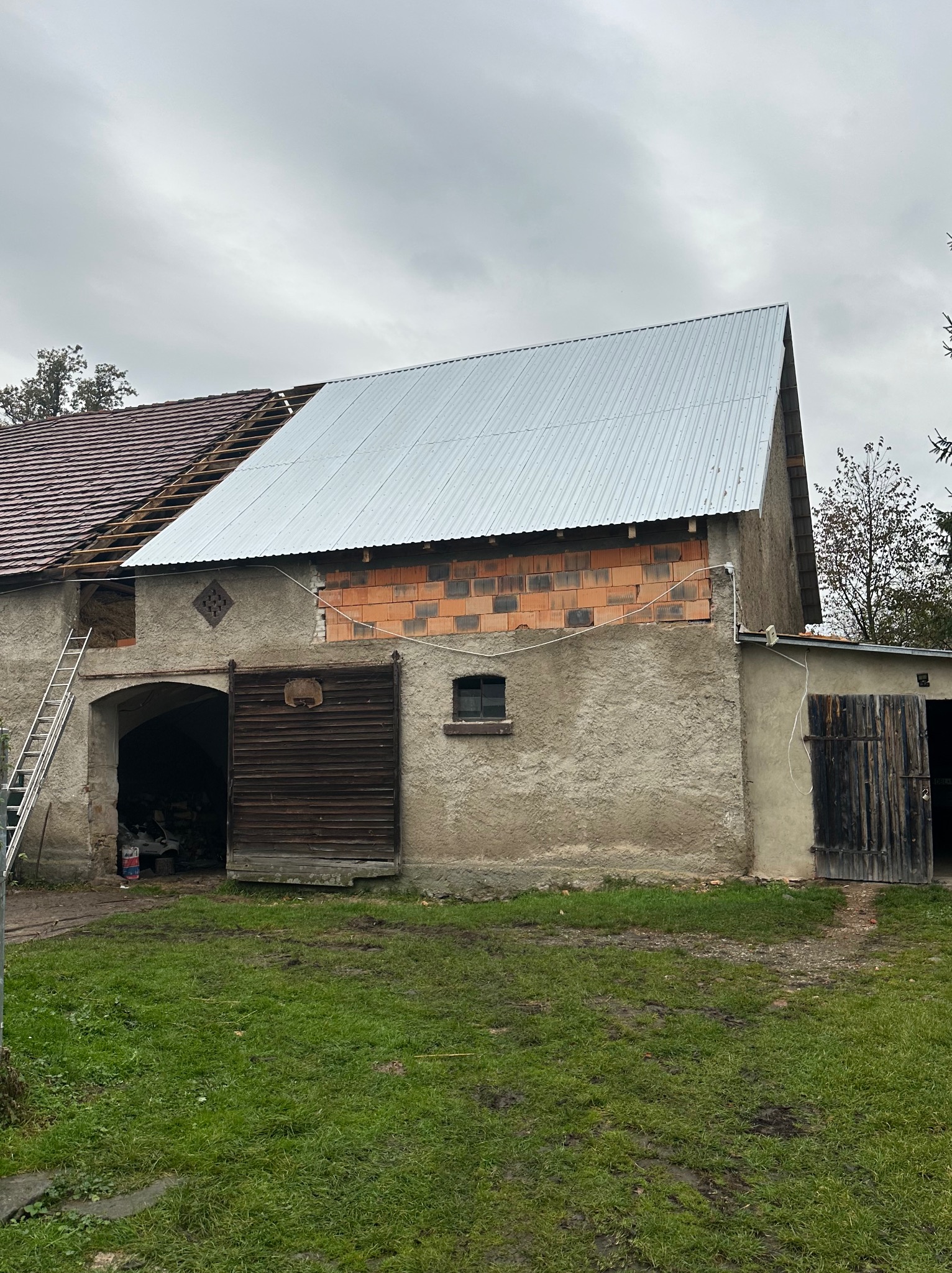 Częściowo wymienione poszycie dachu na budynku gospodarczym. Widoczne cegły i stara elewacja. Drabina oparta o ścianę. Część dachu pokryta blachą falistą.