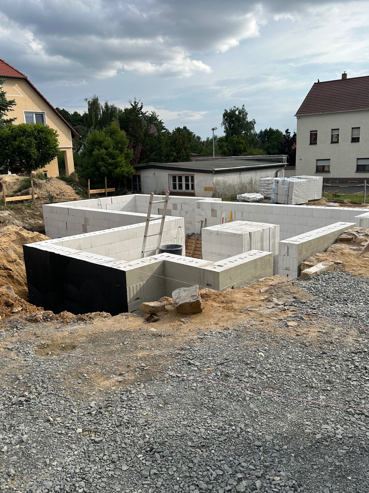 Fundamenty domu z bloczków betonowych, zabezpieczone czarną izolacją, widoczne z ziemi. W tle budynki mieszkalne i pochmurne niebo.