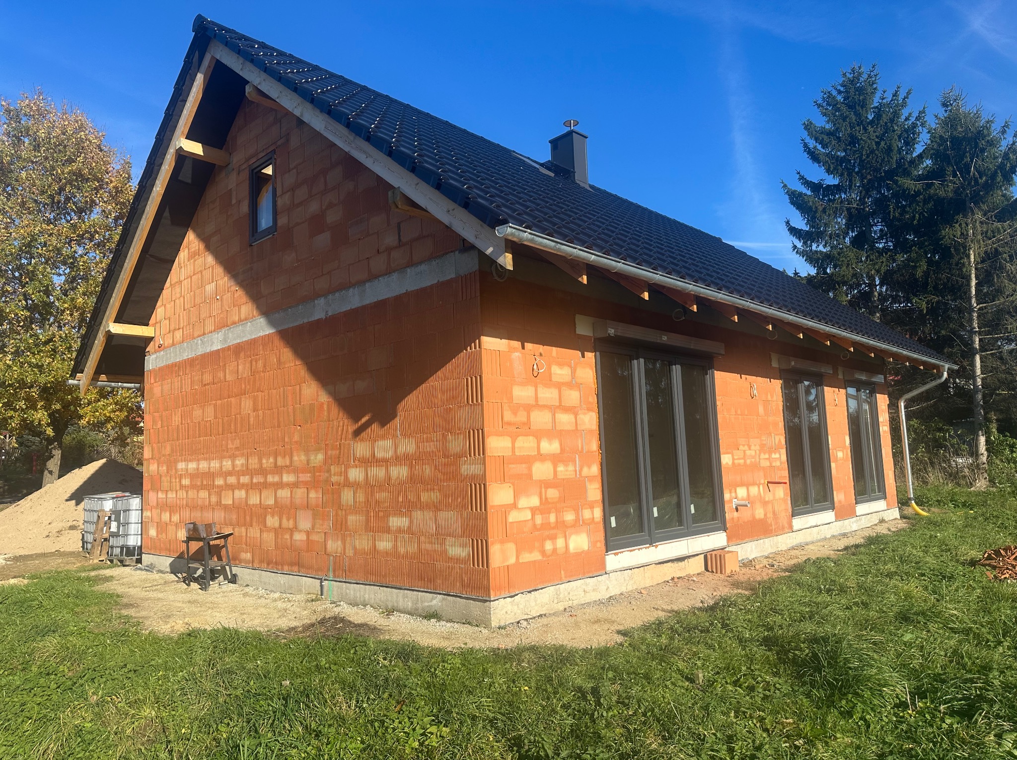 Surowy budynek z czerwonej cegły ceramicznej, z czarnym dachem i szarymi oknami, w trakcie budowy, na tle błękitnego nieba i zielonej trawy.