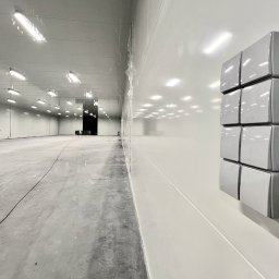 DP spółka z o.o. spółka kom. - Szeroki kadr pustej, jasnej hali przemysłowej z betonową posadzką i białymi ścianami. Na prawej ścianie pionowy panel z ośmioma szarymi modułami sterującymi.
