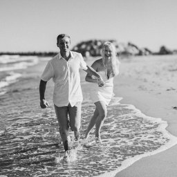 Kinga Wolska Fotografia - Czarno-białe zdjęcie pary biegnącej po plaży, trzymającej się za ręce. Woda obmywa im stopy, a w tle widać zarys lądu. Oboje uśmiechnięci, w lekkich ubraniach.