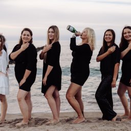 Kinga Wolska Fotografia - Panie na plaży świętują wieczór panieński, jedna pije szampana z butelki, pozostałe w czarnych sukienkach, jedna w bieli z szarfą 'Bride', w tle morze i drewniane pale.