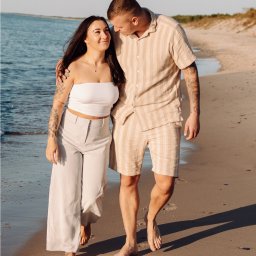 Kinga Wolska Fotografia - Uśmiechnięta para z tatuażami spaceruje brzegiem morza w letnim oświetleniu. Kobieta patrzy na mężczyznę z czułością. Piasek, woda i niebo w tle.