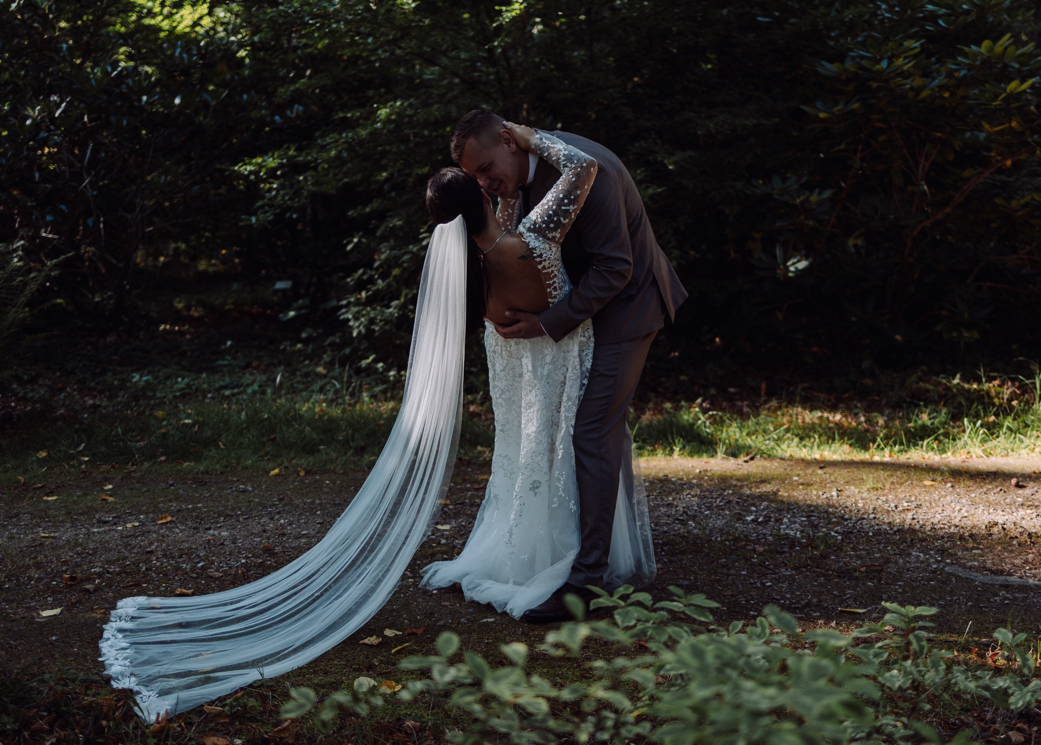 Para młoda w lesie: panna młoda w koronkowej sukni z długim welonem, pan młody w garniturze obejmuje ją, delikatnie pochylając się do pocałunku, drzewa w tle.