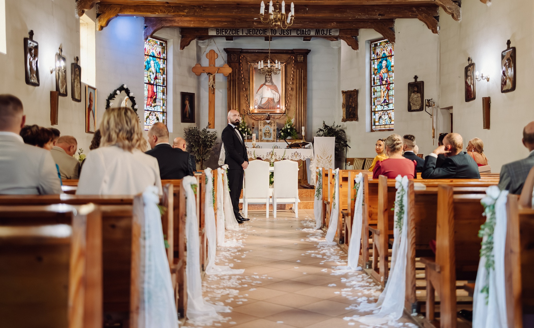 Uroczystość ślubna w kościele: pan młody czeka przy ołtarzu, ławki udekorowane białymi tkaninami i kwiatami, widzowie siedzą na ławkach, witraże w oknach.