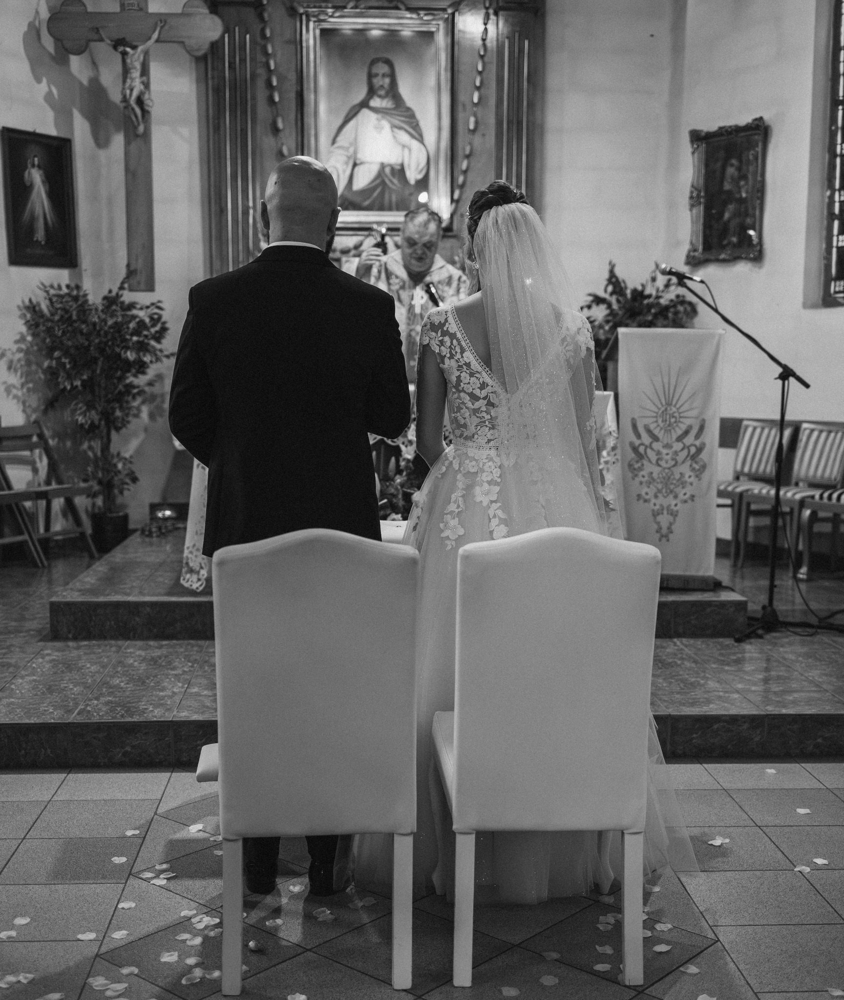 Czarno-białe zdjęcie z ceremonii ślubnej w kościele. Para młoda stoi przed ołtarzem, widoczny ksiądz. Na podłodze rozsypane płatki kwiatów. Ujęcie zza krzeseł.