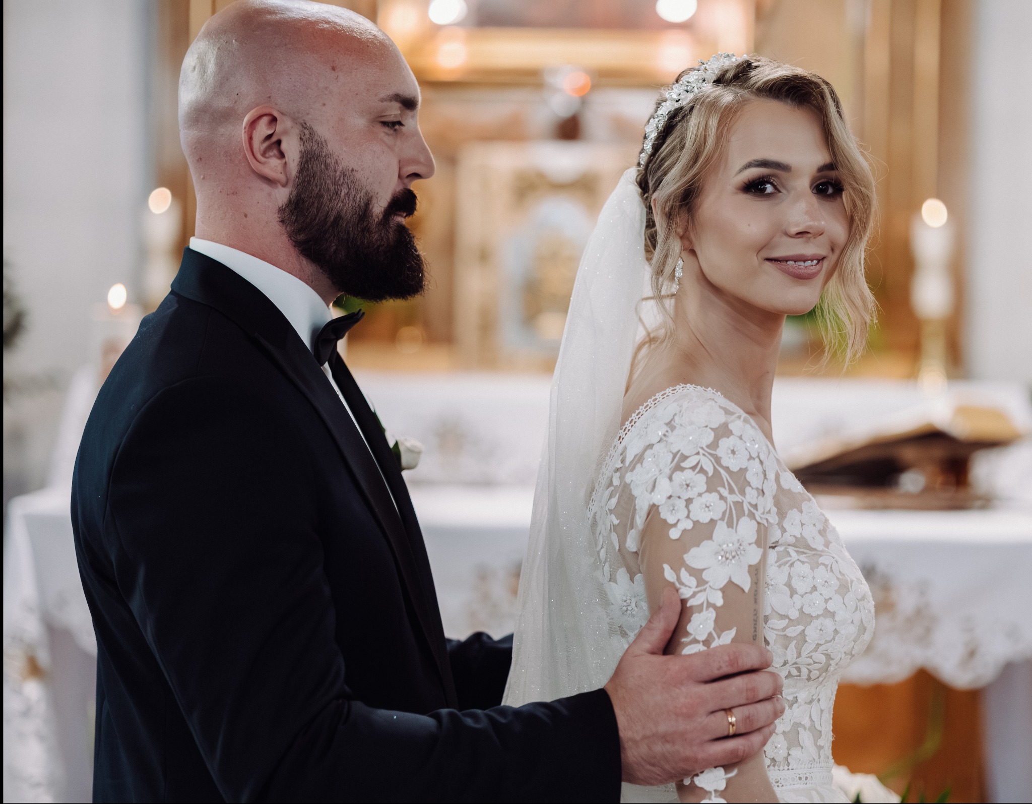 Elegancka para młoda w kościele: panna młoda w koronkowej sukni z welonem, pan młody w czarnym smokingu, delikatne światło, uroczysta atmosfera.