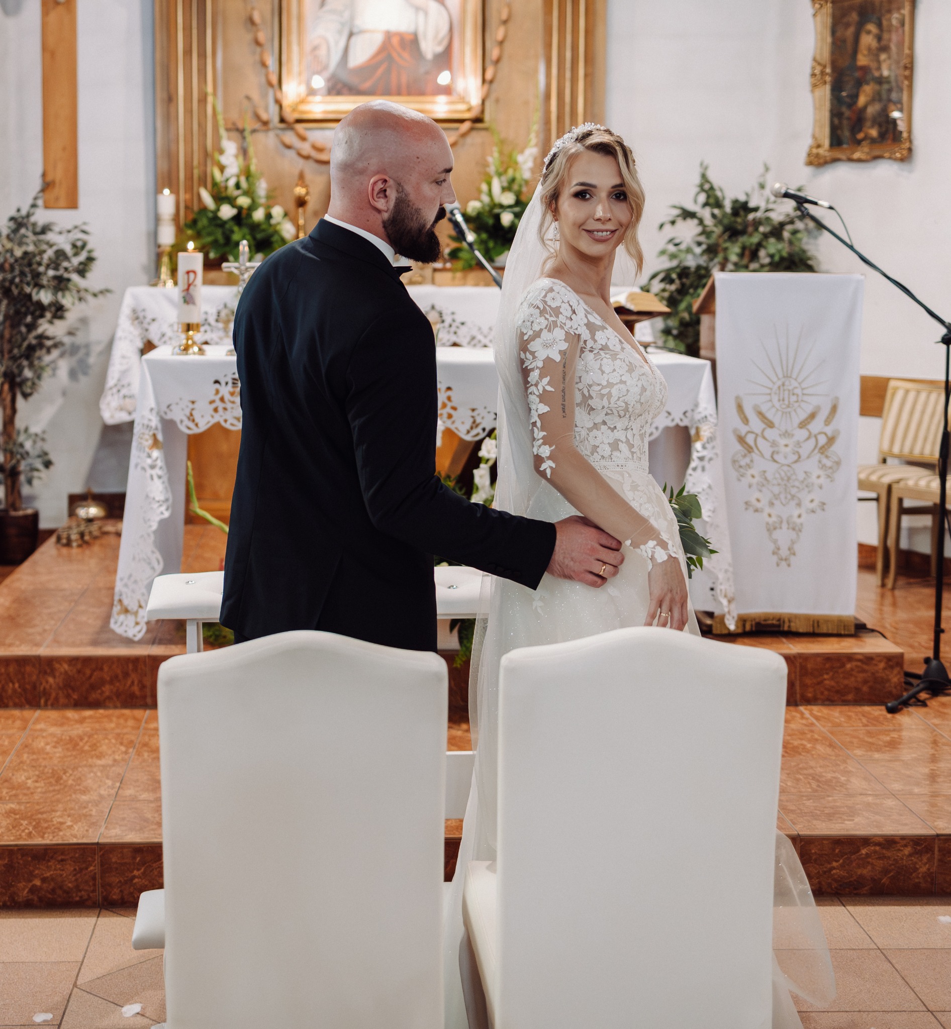 Para młoda w kościele podczas ceremonii ślubnej, panna młoda spogląda w obiektyw, w tle ołtarz i dekoracje kwiatowe, widoczne białe krzesła.