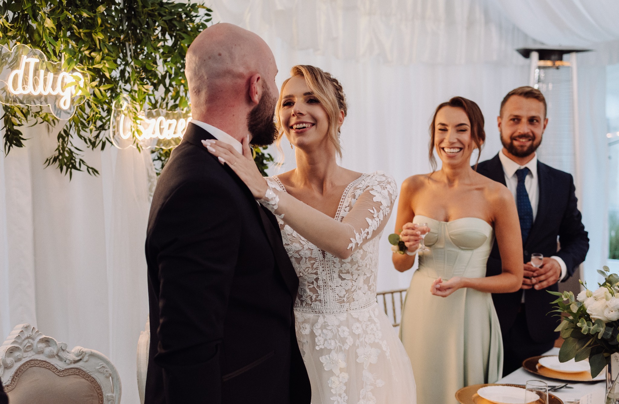 Uśmiechnięta panna młoda w koronkowej sukni dotyka policzka pana młodego na tle pary gości i neonowego napisu 'Długo i szczęśliwie'.