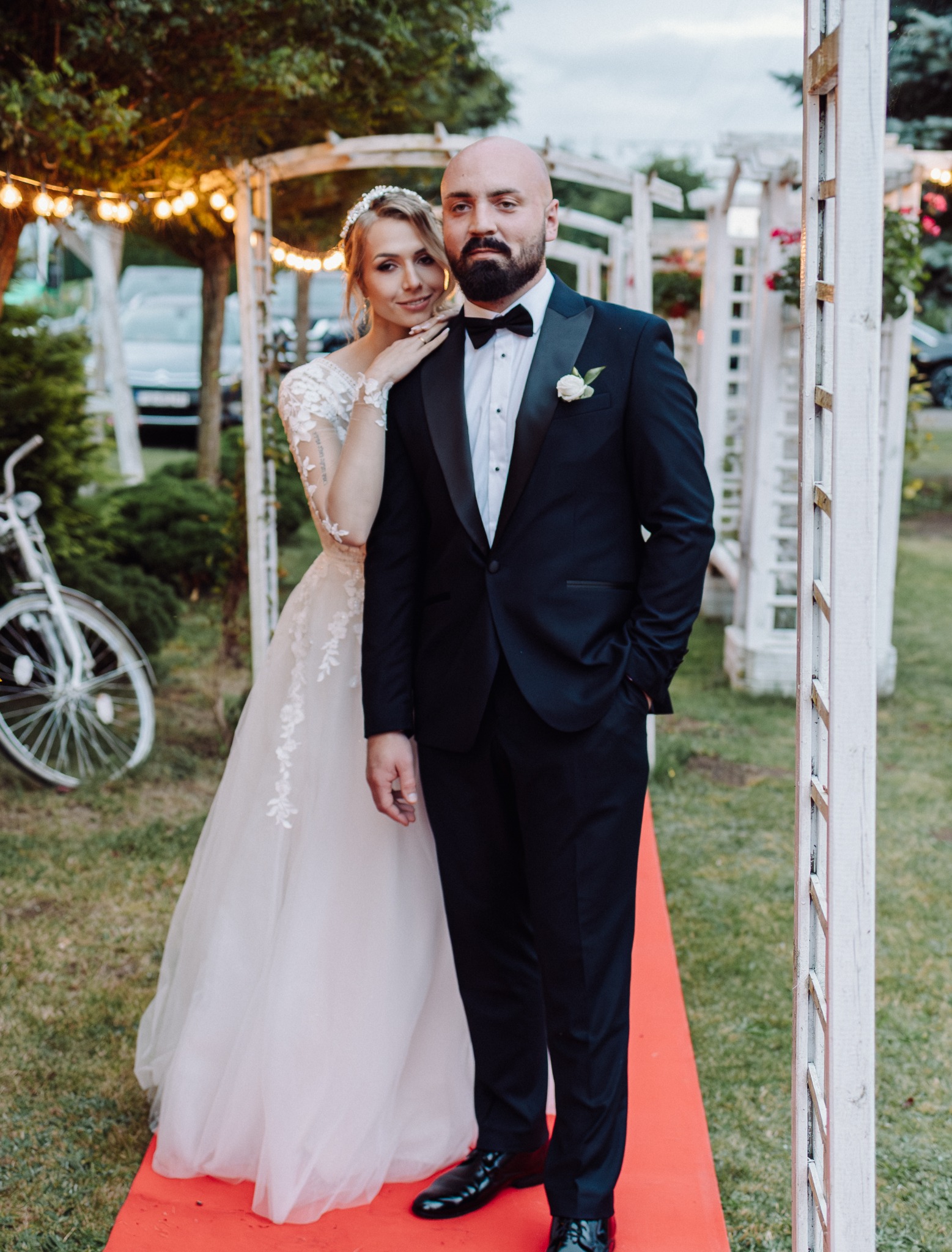 Para młoda w plenerze: panna młoda w koronkowej sukni opiera się o pana młodego w smokingu. W tle białe pergole, czerwony dywan i dekoracyjne oświetlenie.