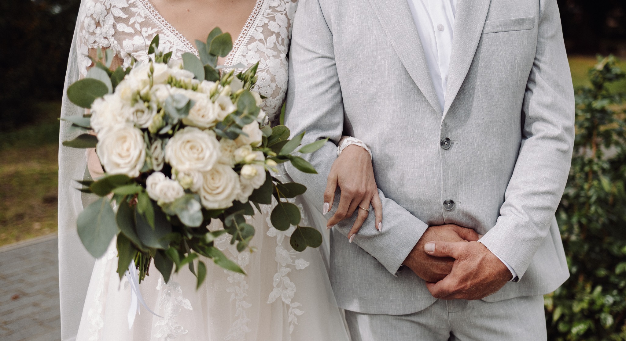 Para młoda: panna młoda w koronkowej sukni trzyma bukiet białych róż, opierając rękę na ramieniu pana młodego w szarym garniturze. Widoczny pierścionek zaręczynowy.