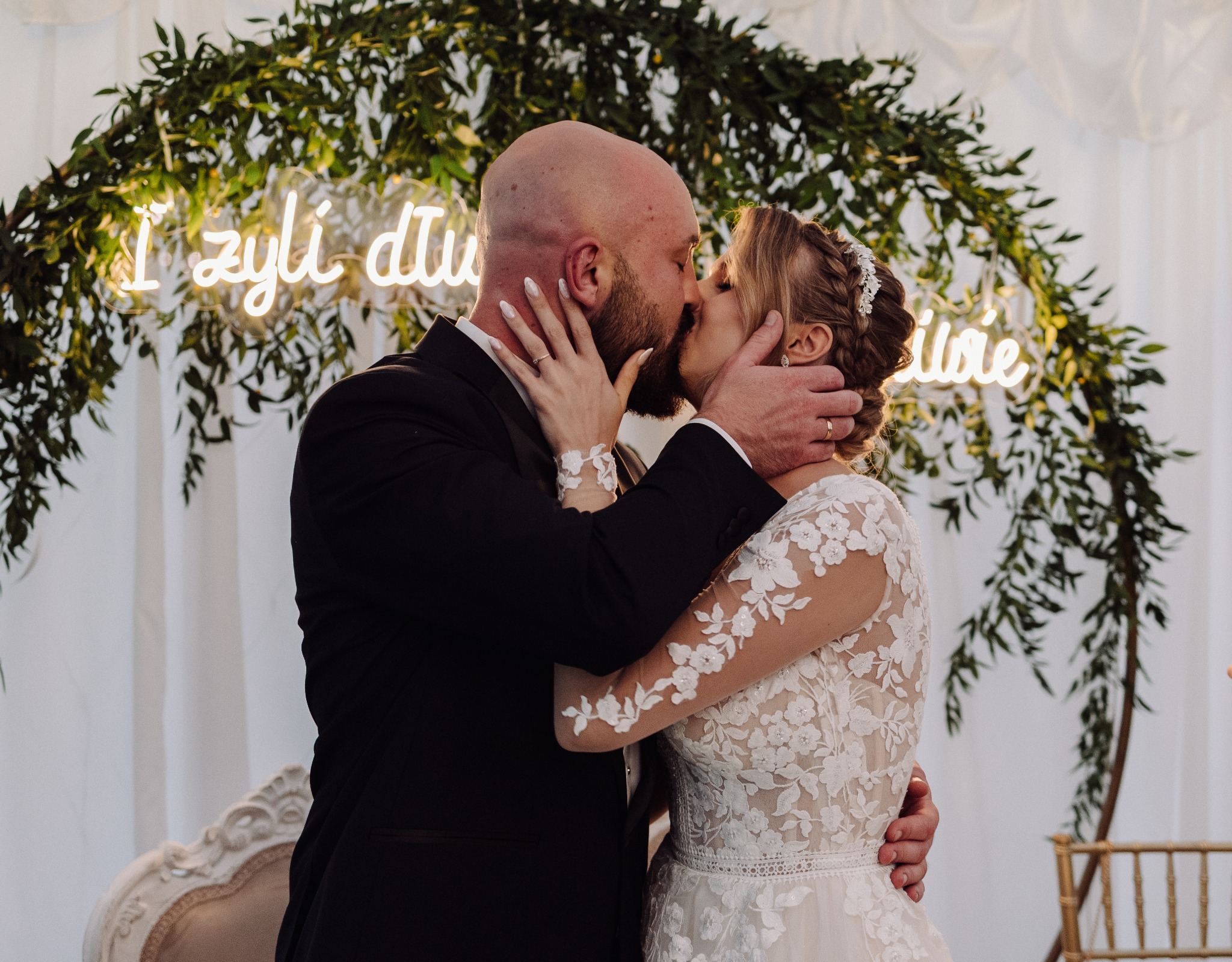 Pocałunek nowożeńców na tle dekoracji z zieleni i neonu. Panna młoda w koronkowej sukni, pan młody w czarnym garniturze. Widoczne obrączki i starannie wykonany makijaż.