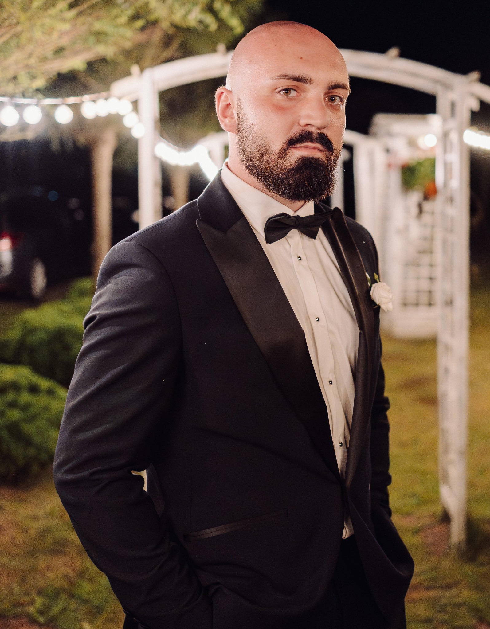 Elegancki mężczyzna w czarnym smokingu i muszce, z przypinką kwiatową, pozuje na tle dekoracji ślubnej z oświetleniem.