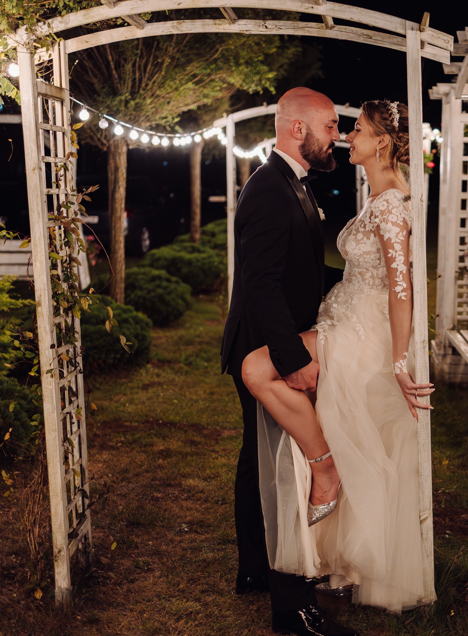 Para młoda w ogrodowej altanie, oświetlonej girlandami. Panna młoda w sukni z koronkowymi aplikacjami opiera się o konstrukcję, unosząc nogę. Pan młody w smokingu.