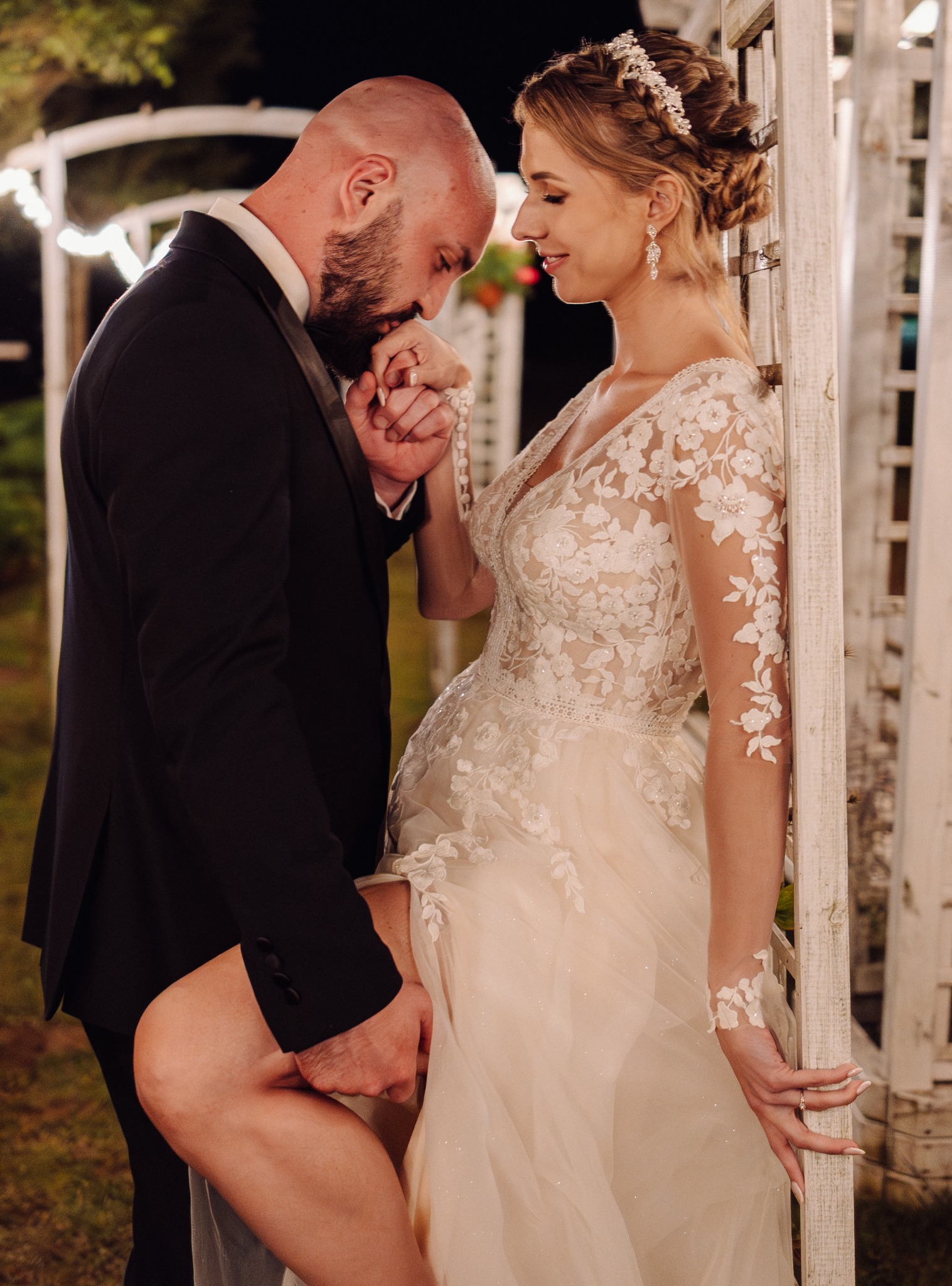 Elegancka para młoda: mężczyzna w smokingu całuje dłoń kobiety w koronkowej sukni, opartą o białą altanę z kwiatami, oświetloną lampkami.