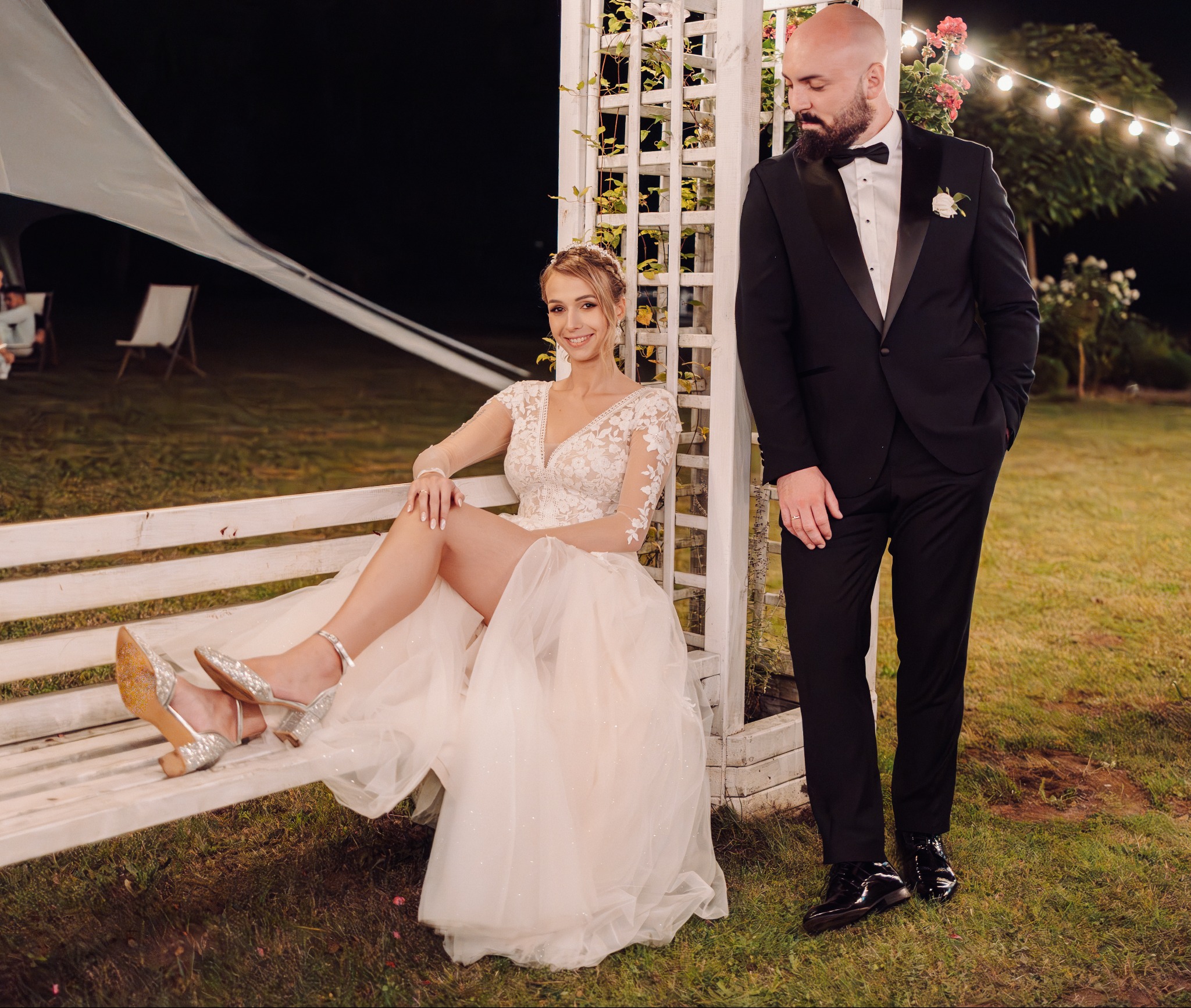 Uśmiechnięta panna młoda w koronkowej sukni i srebrnych butach siedzi na białej ławce, obok stoi pan młody w smokingu. Romantyczna sesja ślubna w Kawno.