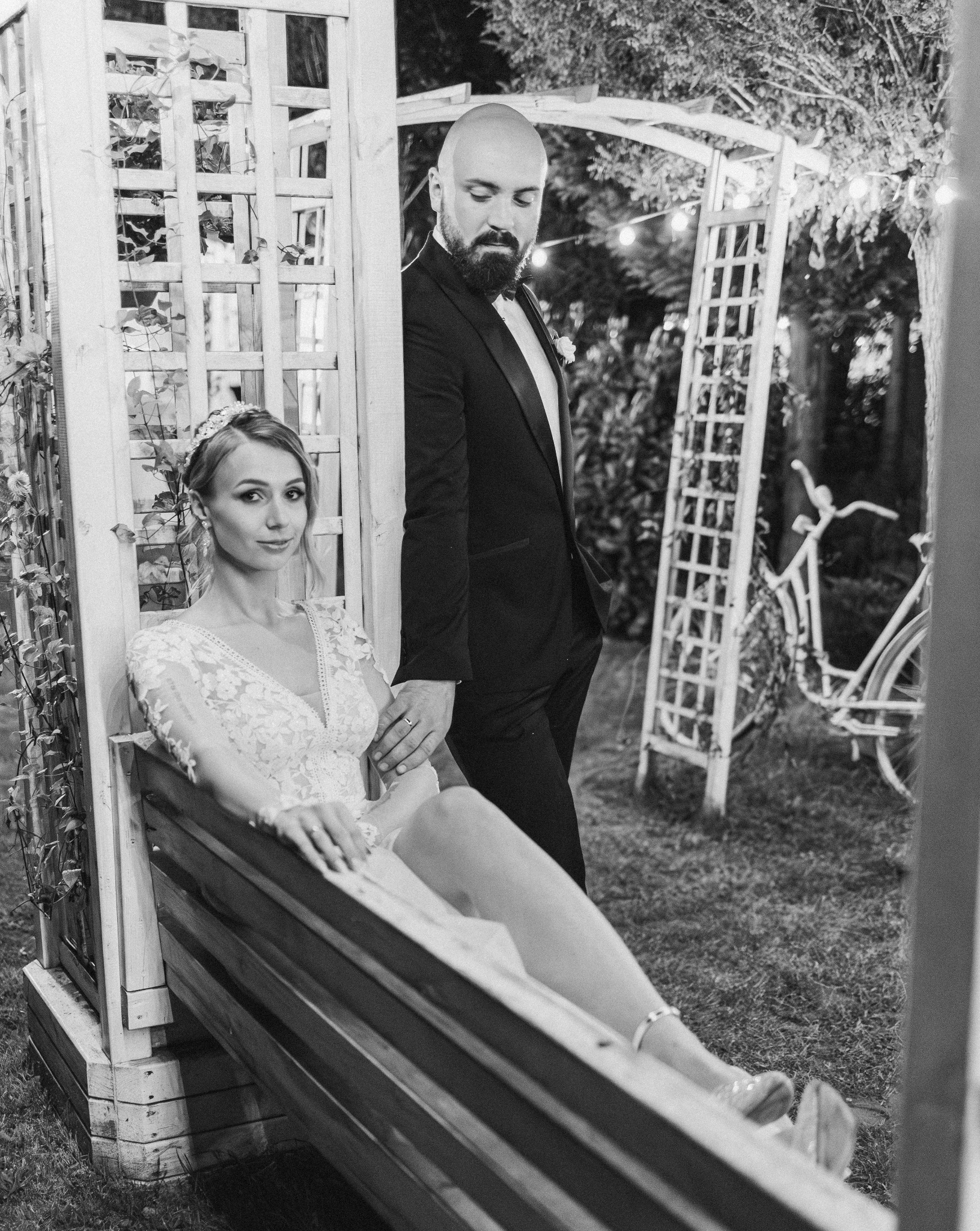 Czarno-białe zdjęcie pary młodej: panna młoda w koronkowej sukni siedzi na ławce, pan młody w smokingu stoi obok, trzymając ją za rękę, w tle dekoracje i rower.