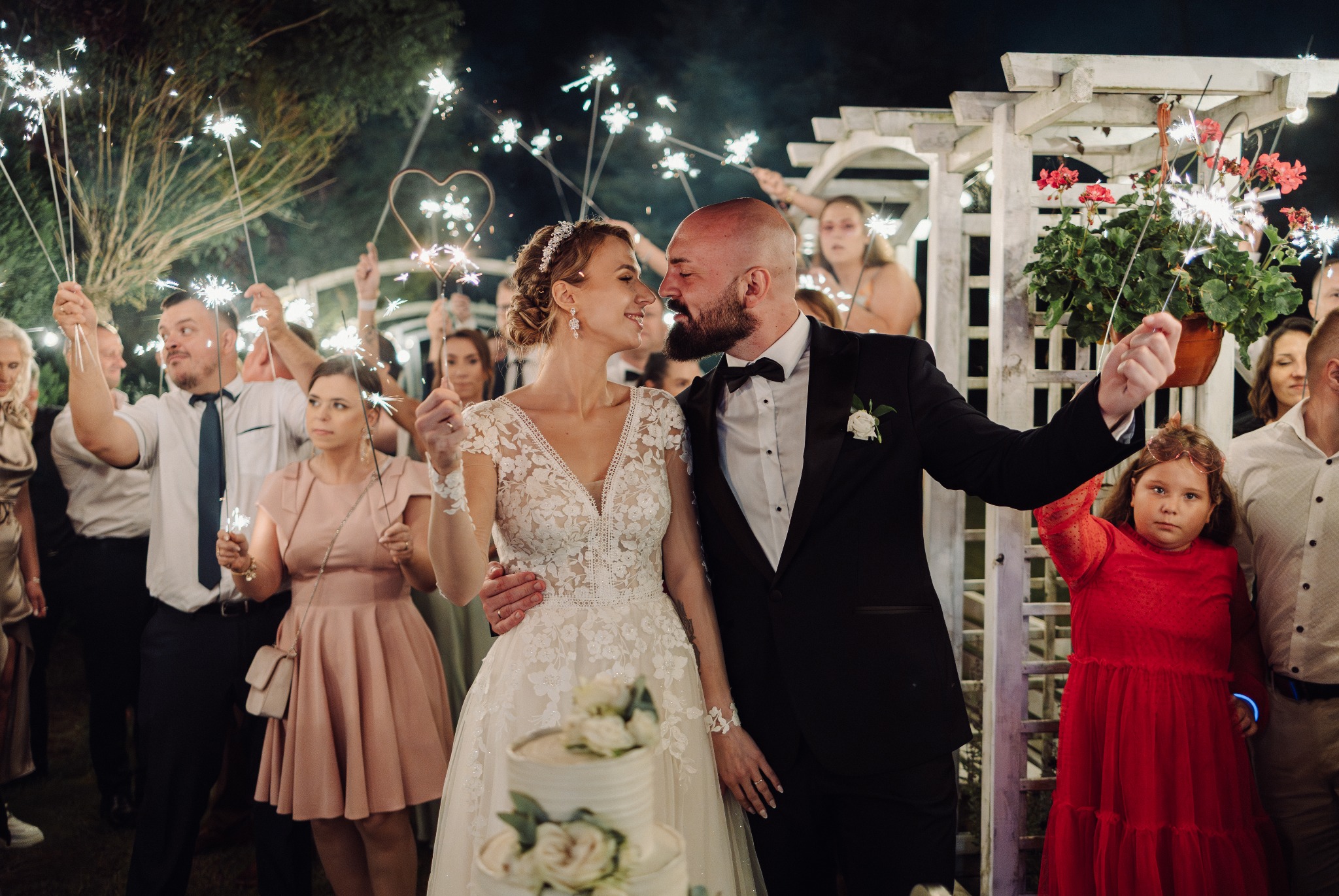 Uśmiechnięta para młoda w otoczeniu gości z zimnymi ogniami, w tle biała pergola i kwiaty. Panna młoda w koronkowej sukni, pan młody w smokingu.