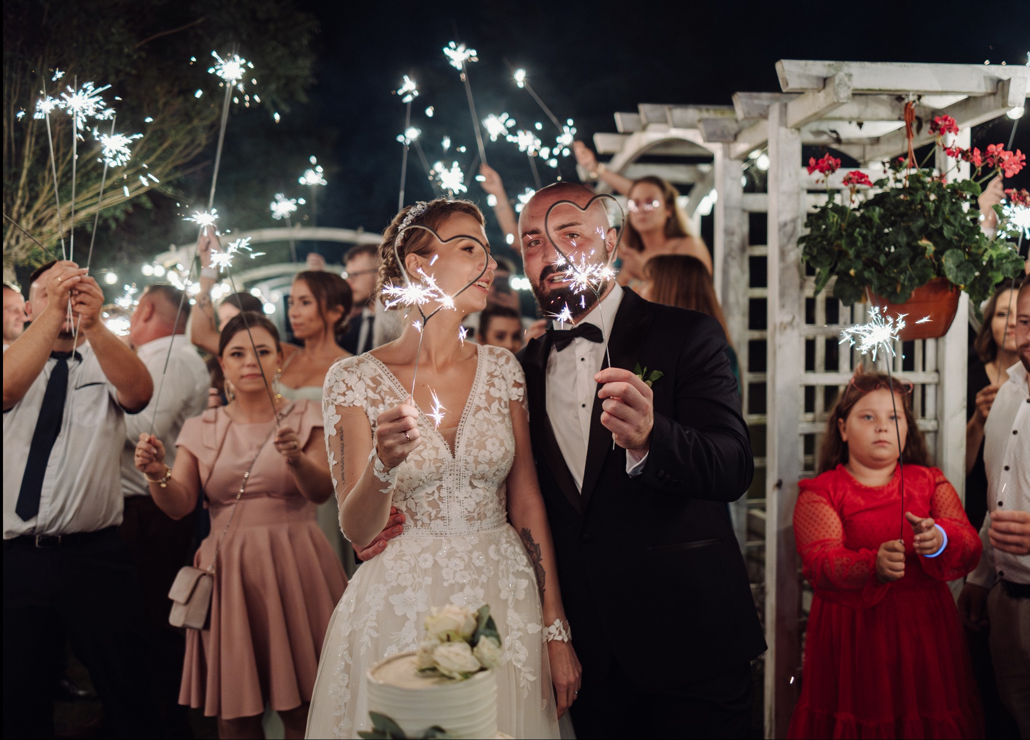 Uśmiechnięta para młoda trzymająca zimne ognie w kształcie serc, otoczona gośćmi z zimnymi ogniami na tle białej altany i zieleni. Romantyczna atmosfera.