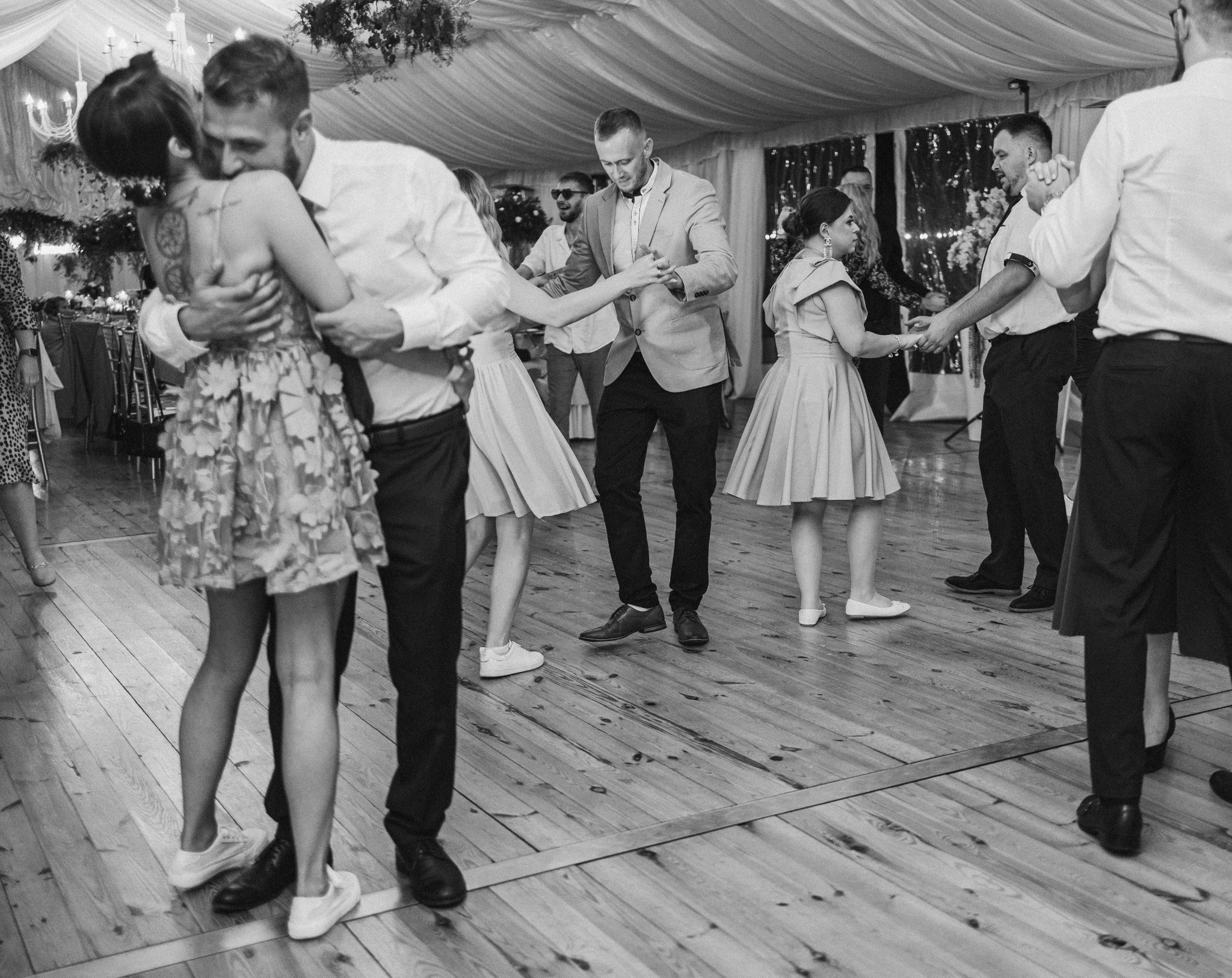 Czarno-białe zdjęcie z wesela: pary tańczą na parkiecie, oświetlonym światłem żyrandoli. Pan młody obejmuje pannę młodą, w tle goście w ruchu.