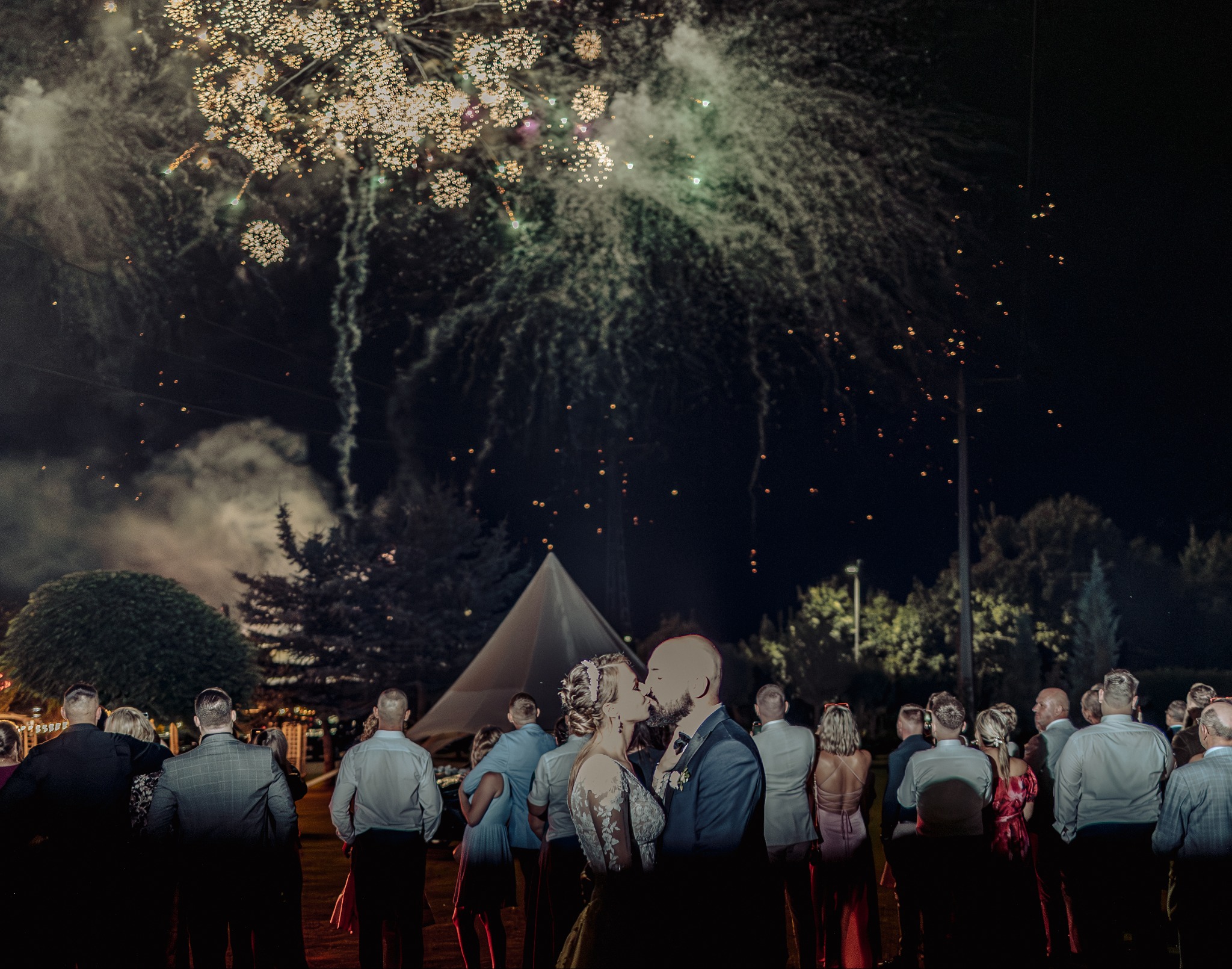 Pocałunek pary młodej na tle nocnego nieba rozświetlonego fajerwerkami, otoczonej gośćmi weselnymi i namiotem tipi w tle. Romantyczna fotografia ślubna.