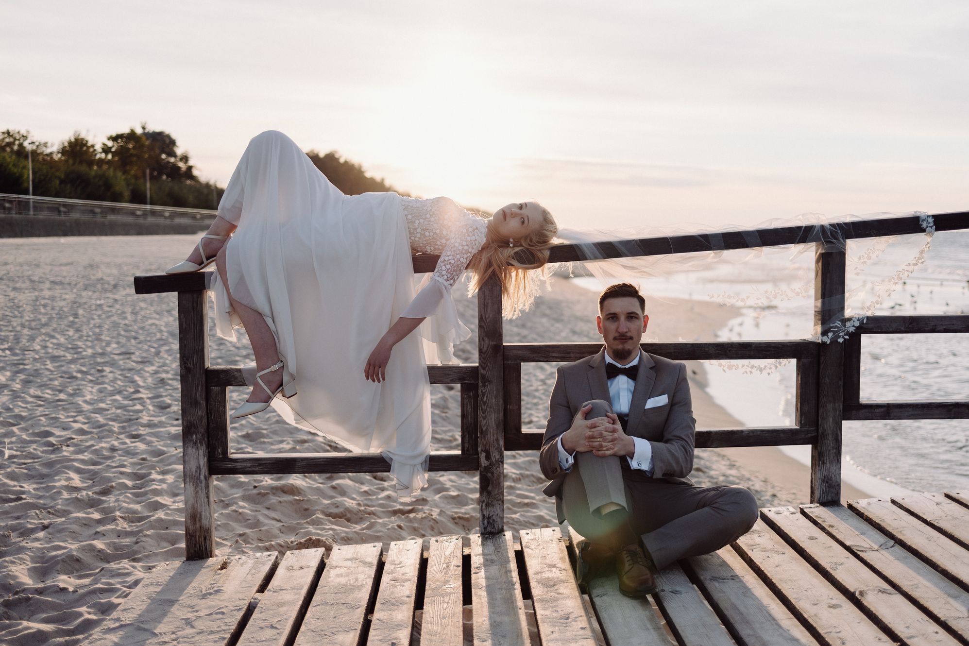 Para młoda na drewnianym pomoście: panna młoda w białej sukni leży na barierce, pan młody w garniturze siedzi obok. Romantyczna sesja ślubna na plaży o zachodzie słońca.