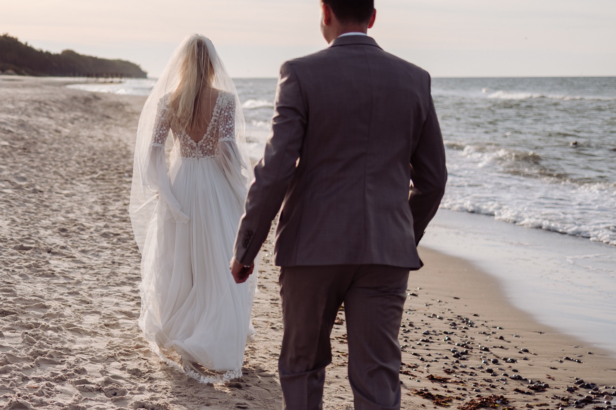 Para młoda spaceruje brzegiem morza. Panna młoda w zwiewnej sukni z koronkowymi plecami i długim welonem, pan młody w szarym garniturze. Romantyczna sceneria, delikatne światło.