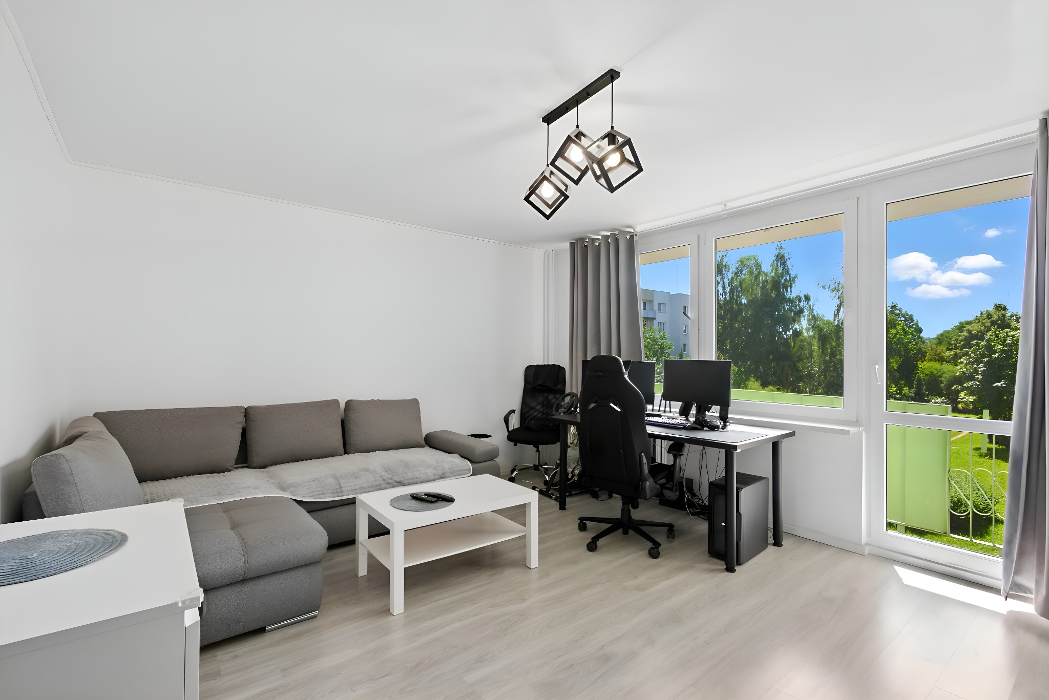 Jasne wnętrze pokoju z szarą sofą, białym stolikiem kawowym i biurkiem z komputerami pod oknem z widokiem na zieleń. Nowoczesna lampa sufitowa.