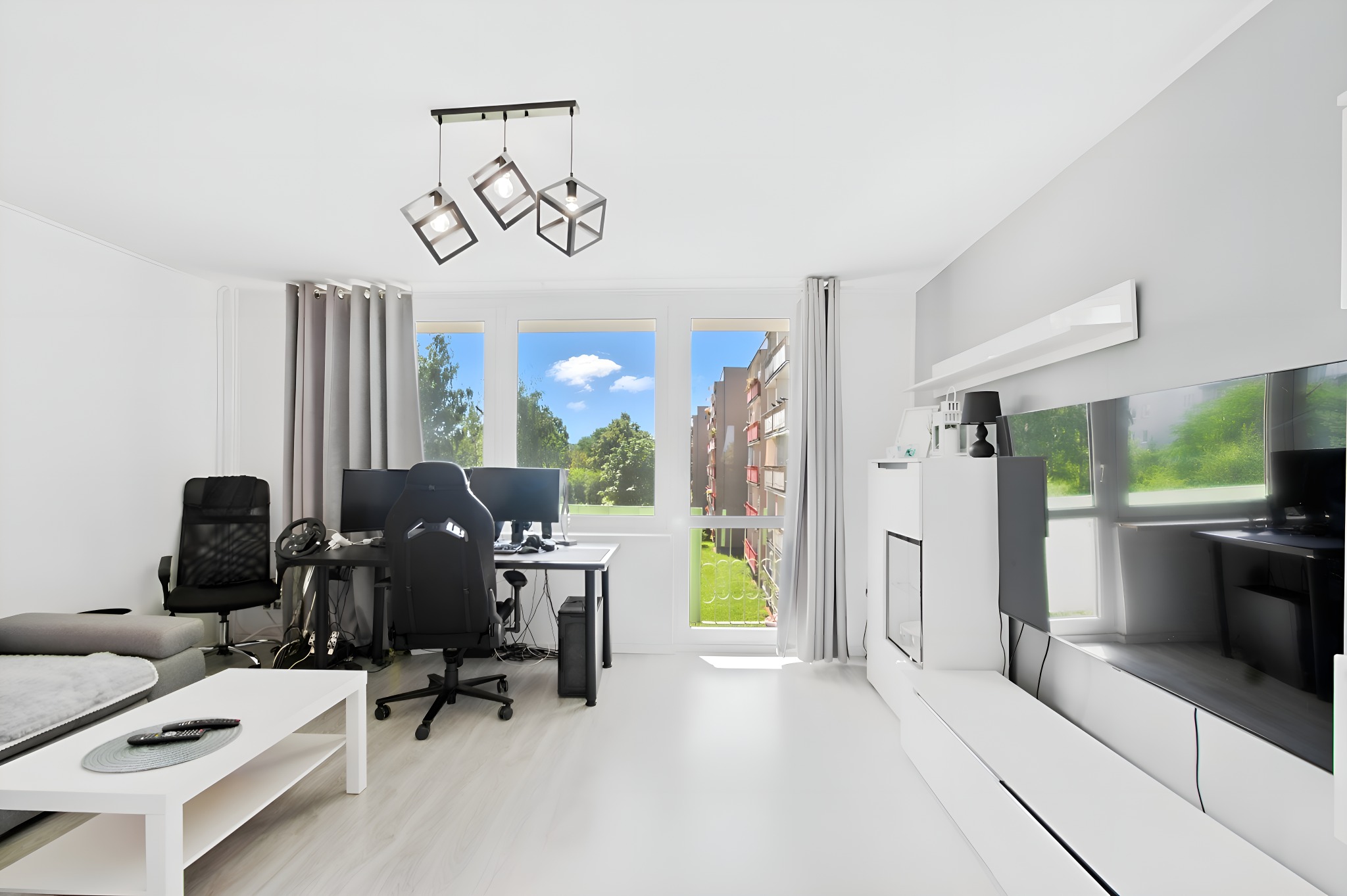Jasne, przestronne wnętrze mieszkania z biurkiem gamingowym i widokiem na zieleń za oknem. Minimalistyczny design, białe meble i podłogi, szare zasłony. Nowoczesne oświetlenie.