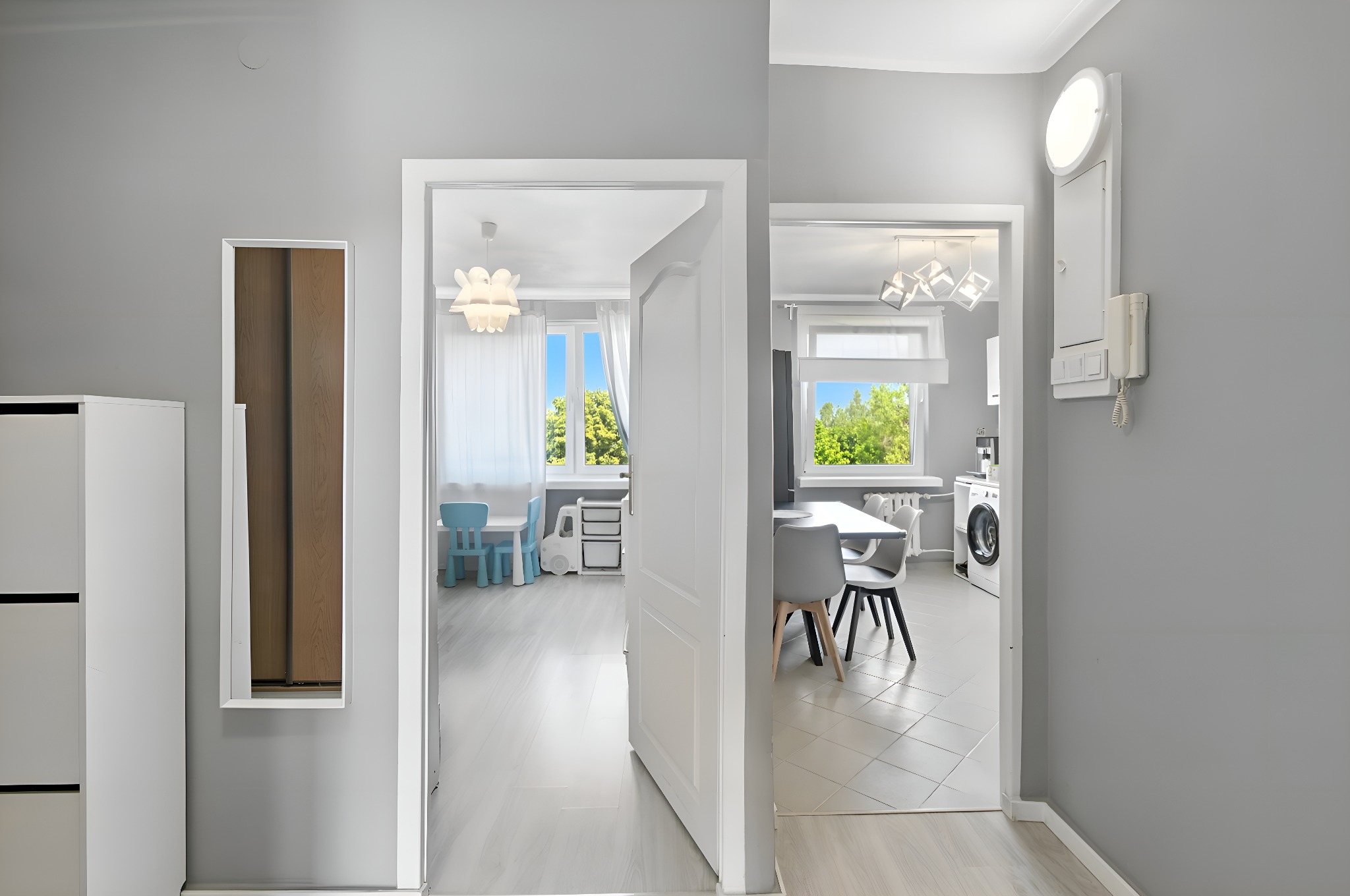 Jasne, przestronne wnętrze mieszkania z widokiem na pokój dziecięcy i kuchnię przez otwarte, białe drzwi. Szare ściany, białe meble, naturalne światło.