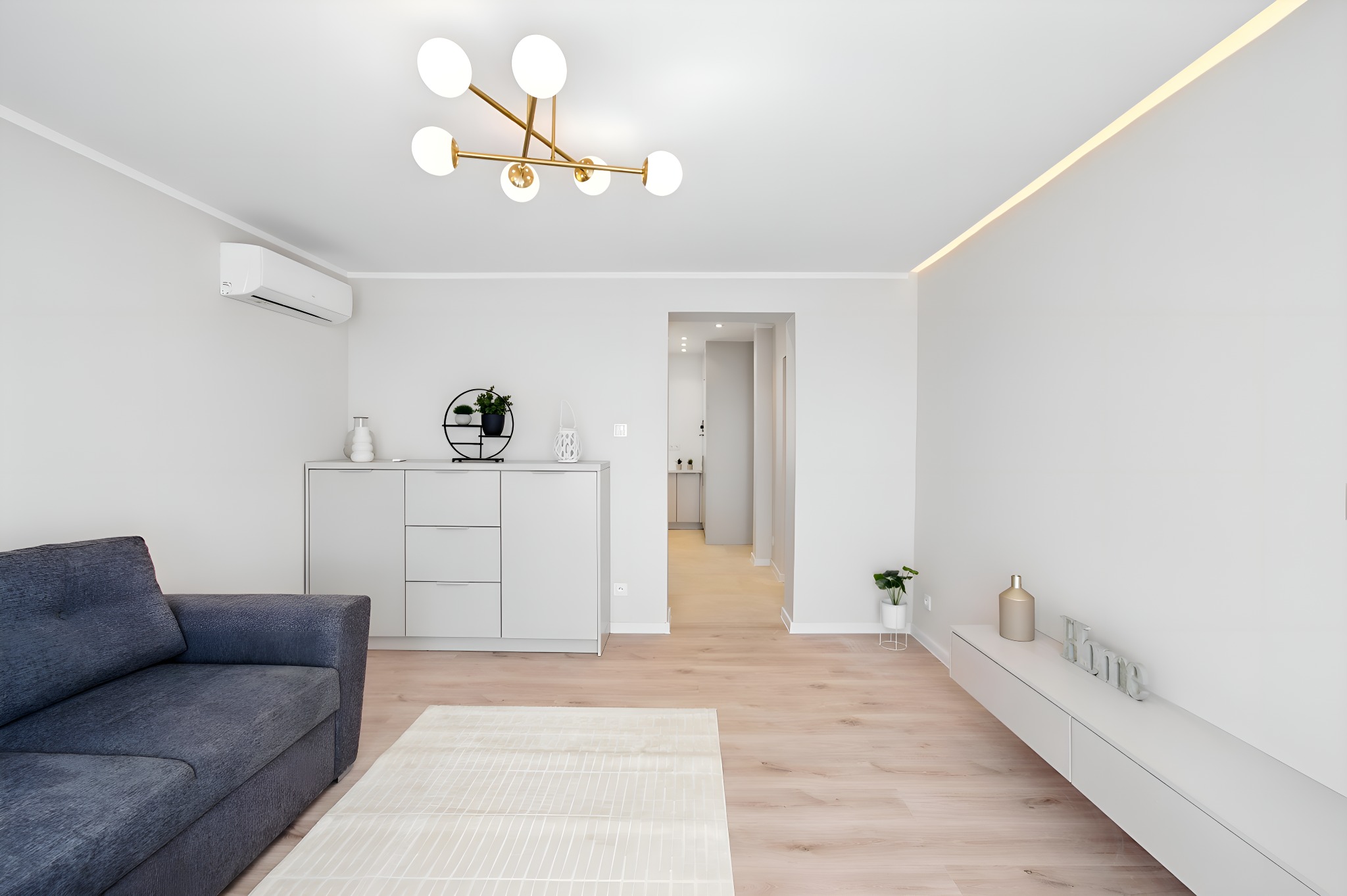 Jasne, przestronne wnętrze z szarą sofą, białymi meblami i złotą lampą sufitową. Minimalistyczny design z drewnianą podłogą i dekoracjami roślinnymi.