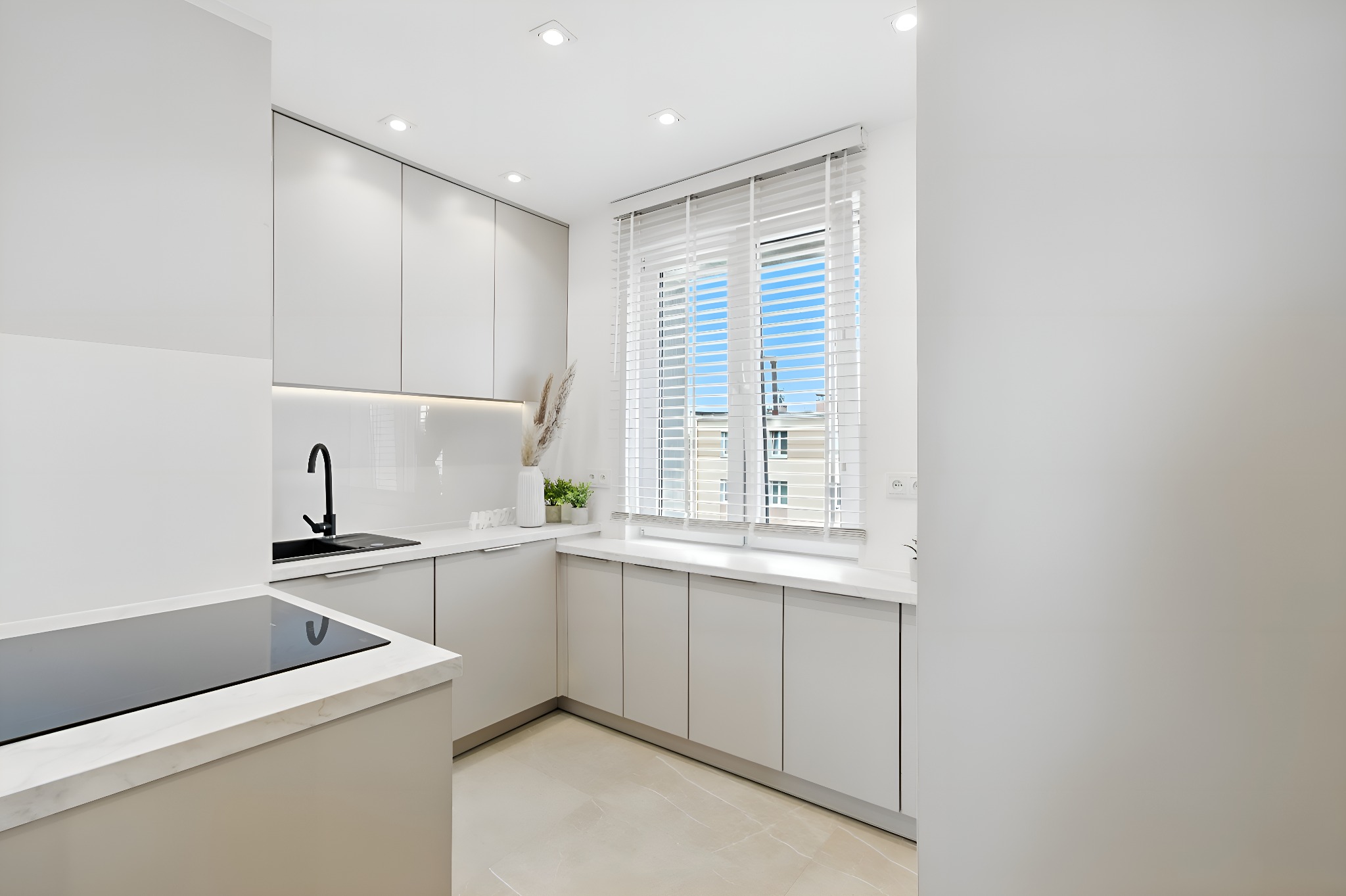 Jasna, nowoczesna kuchnia z białymi szafkami, czarną armaturą i marmurowym blatem. Okno z żaluzjami w tle wpuszcza naturalne światło.