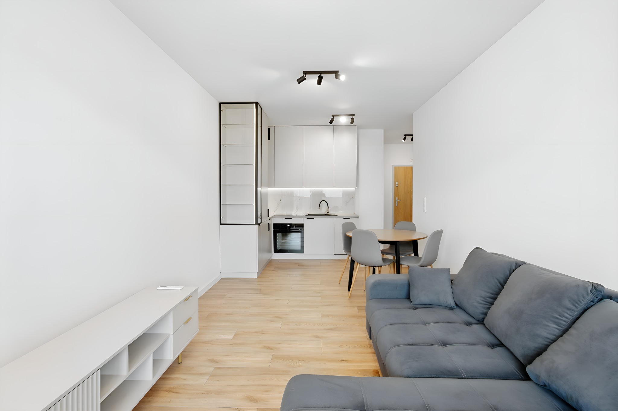 Jasne wnętrze mieszkania z szarą sofą, drewnianą podłogą i minimalistyczną białą kuchnią z czarnymi akcentami. Nowoczesne oświetlenie punktowe.