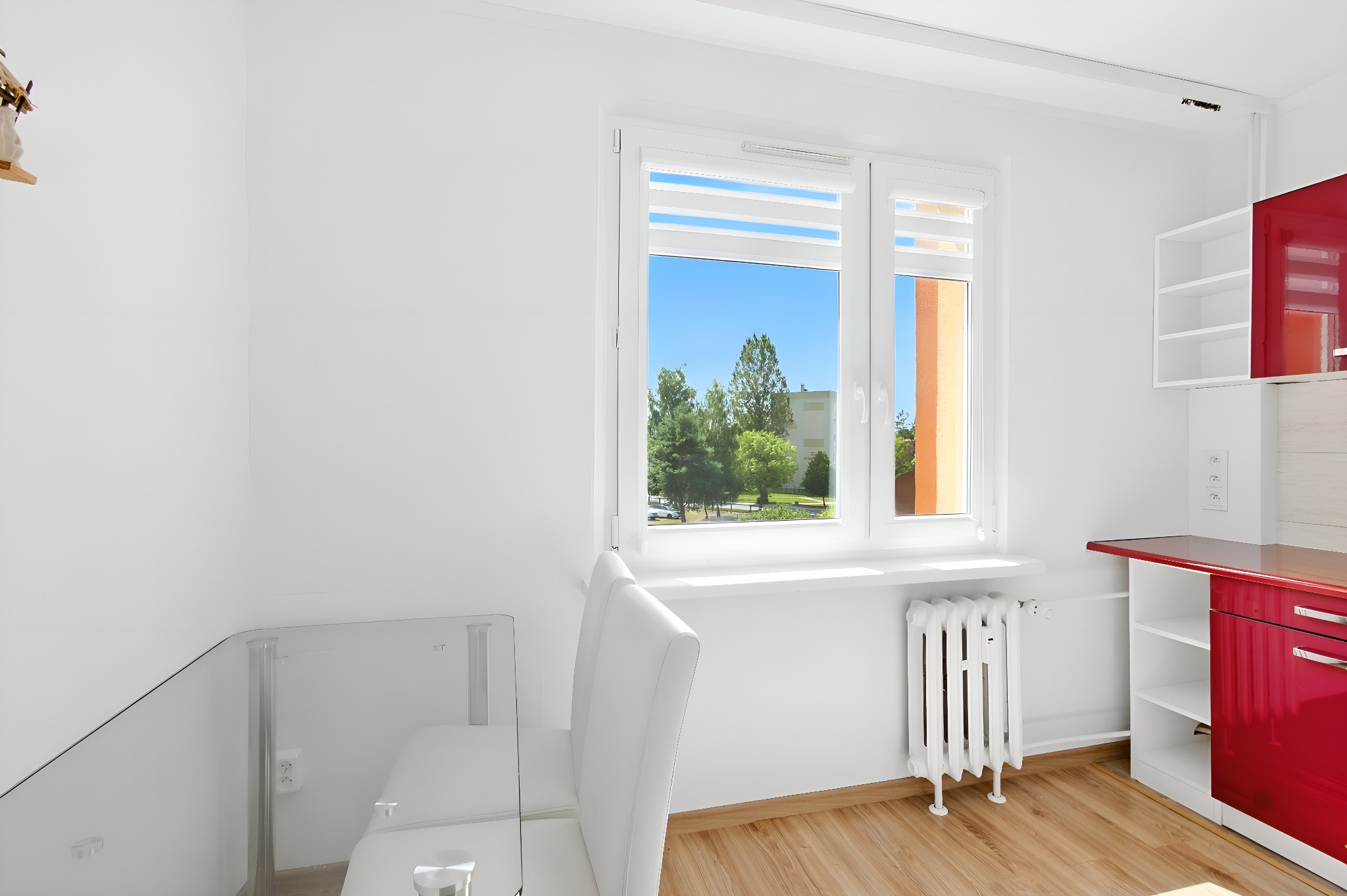 Jasna kuchnia z oknem na zieleń. Białe krzesła i szklany stół. Czerwone szafki. Grzejnik pod oknem. Parkiet na podłodze. Minimalistyczny wystrój.