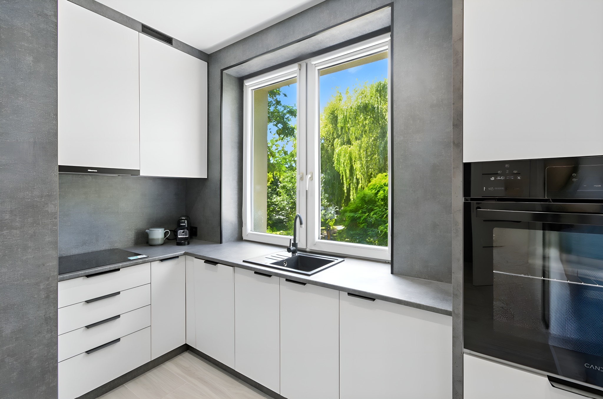 Nowoczesna kuchnia z białymi szafkami i szarymi ścianami. Duże okno z widokiem na zieleń. Czarny zlew i bateria. Minimalistyczny design.